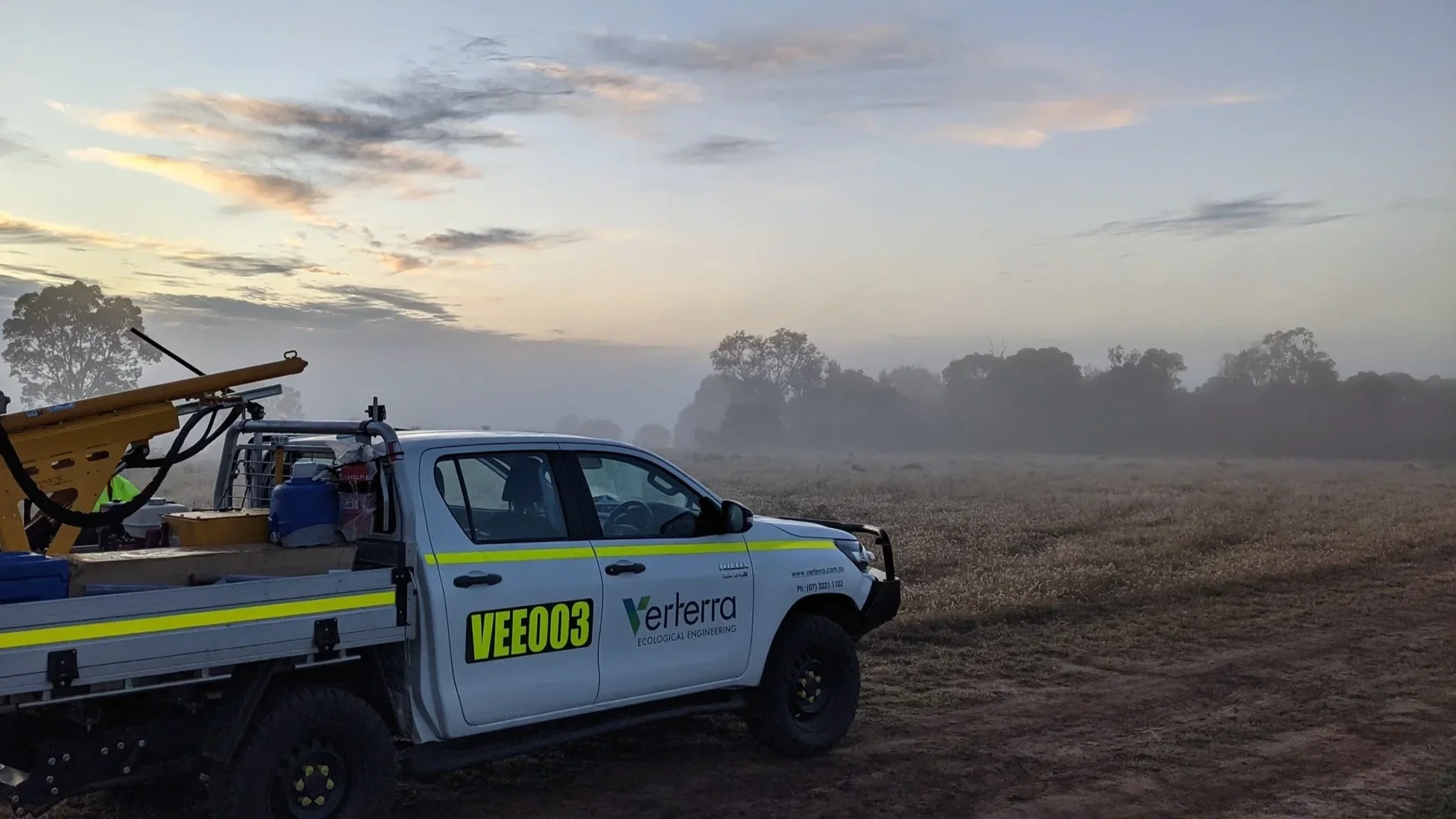 A utility truck with equipment parked on a field during dawn or dusk, with trees and cloudy sky in the background.