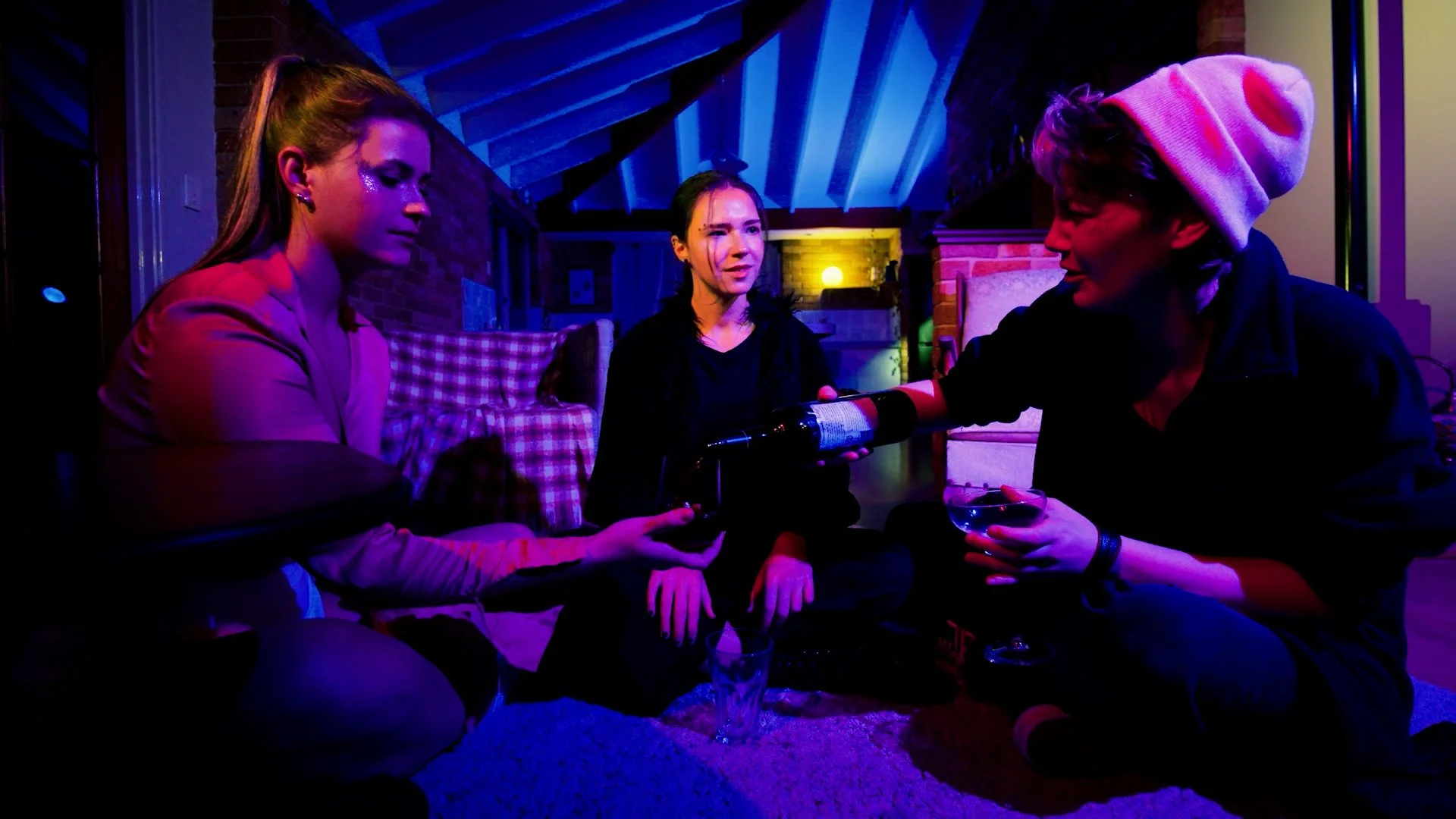 Three women sitting on the floor in a dimly lit room with colorful lighting, one holding a bottle and another with a wine glass, engaged in conversation.