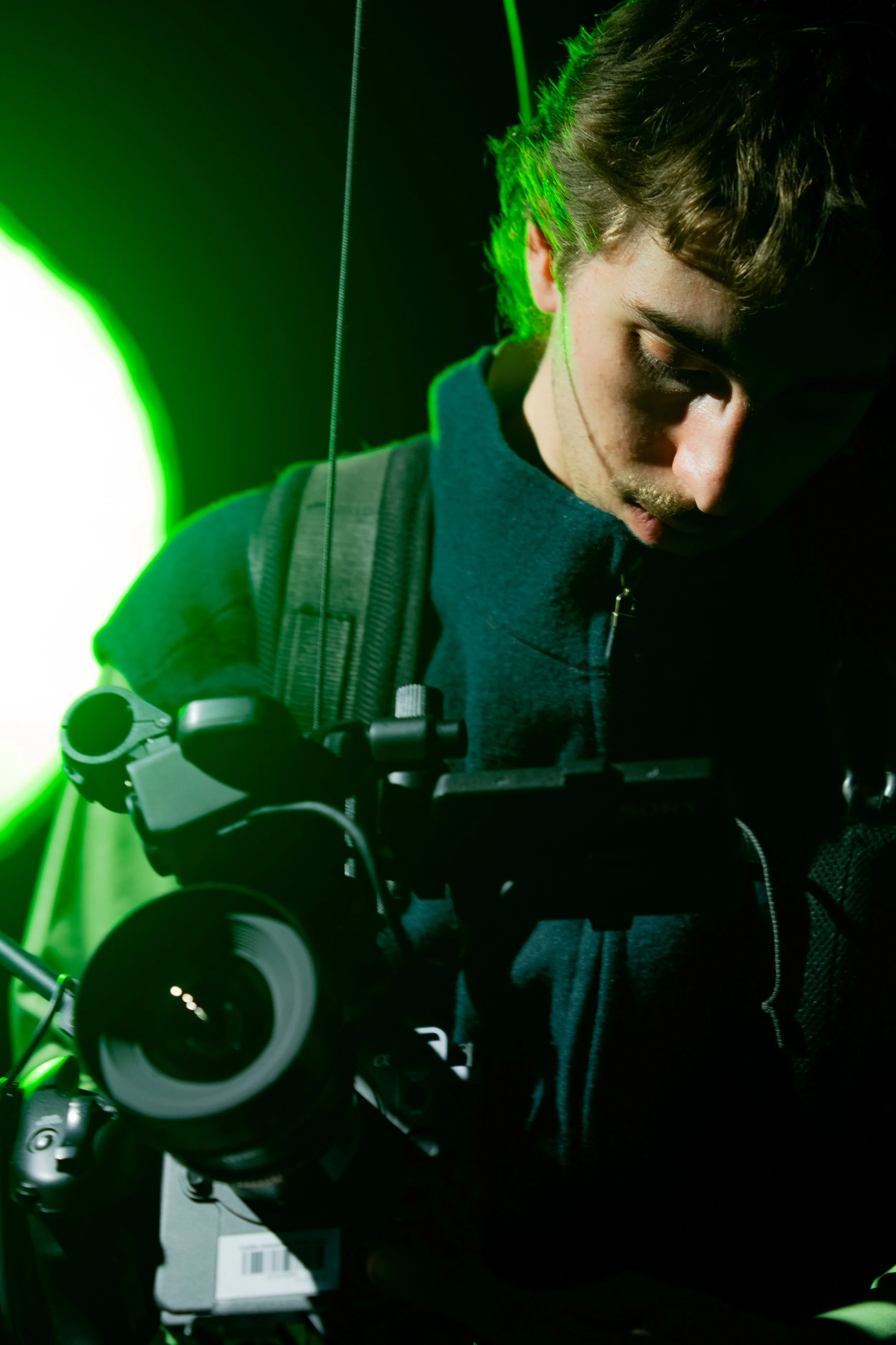 A young man with light brown hair, wearing a dark hoodie and a backpack, operating a professional camera with a large lens, illuminated by green lighting.