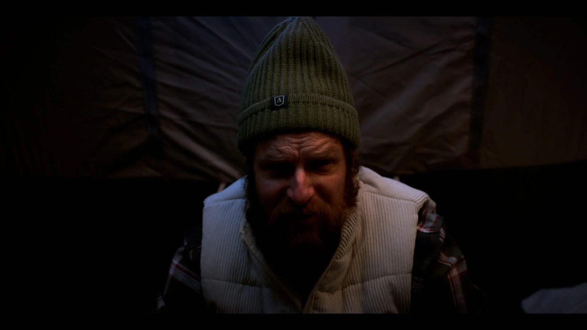 A man with a red beard wearing a green knit beanie and a beige vest over a plaid shirt looks downward with a serious expression.