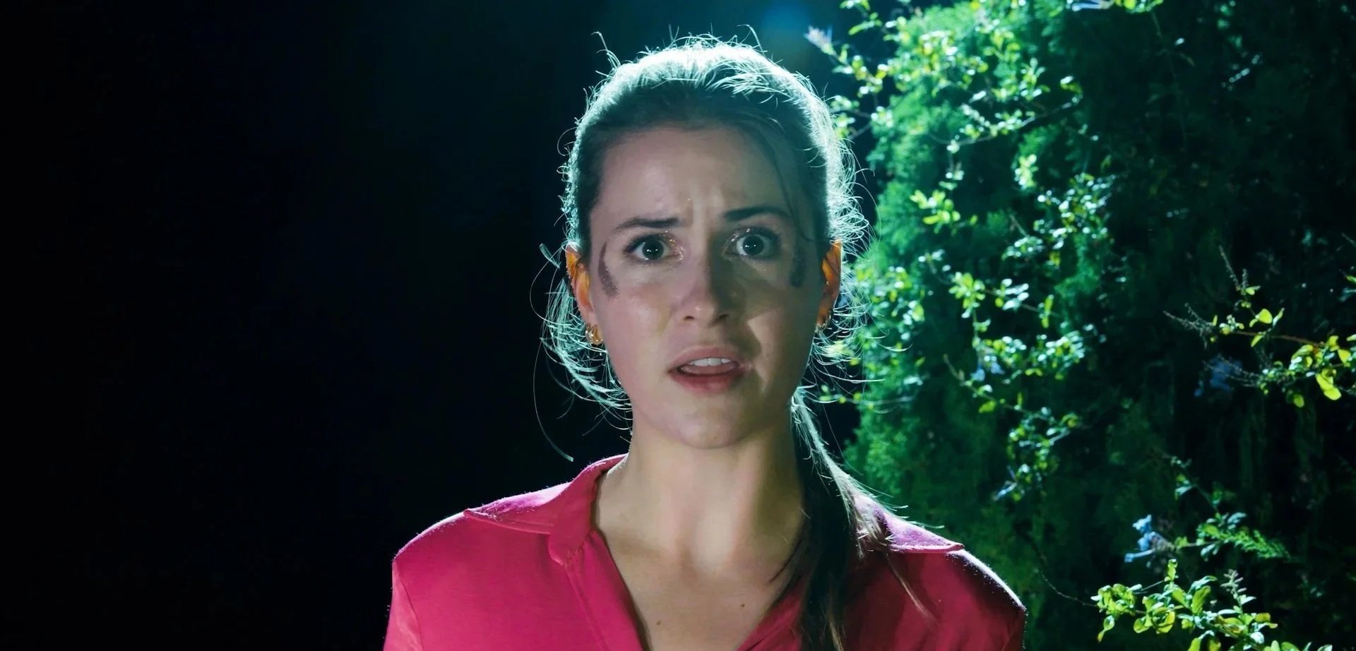 A woman with long dark hair tied back, wearing a pink shirt, has a surprised or shocked expression with dirt or mud on her face, standing outdoors with green foliage in the background and sunlight filtering through the leaves.