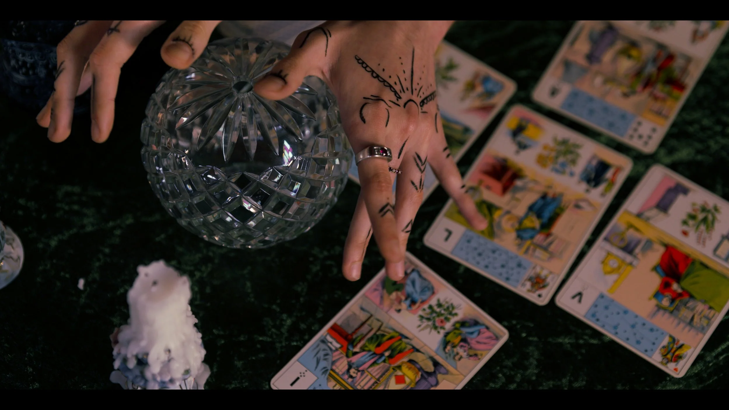 A hand with tattoos and rings holding a faceted glass object, next to tarot cards spread out on a dark surface, with candles nearby.