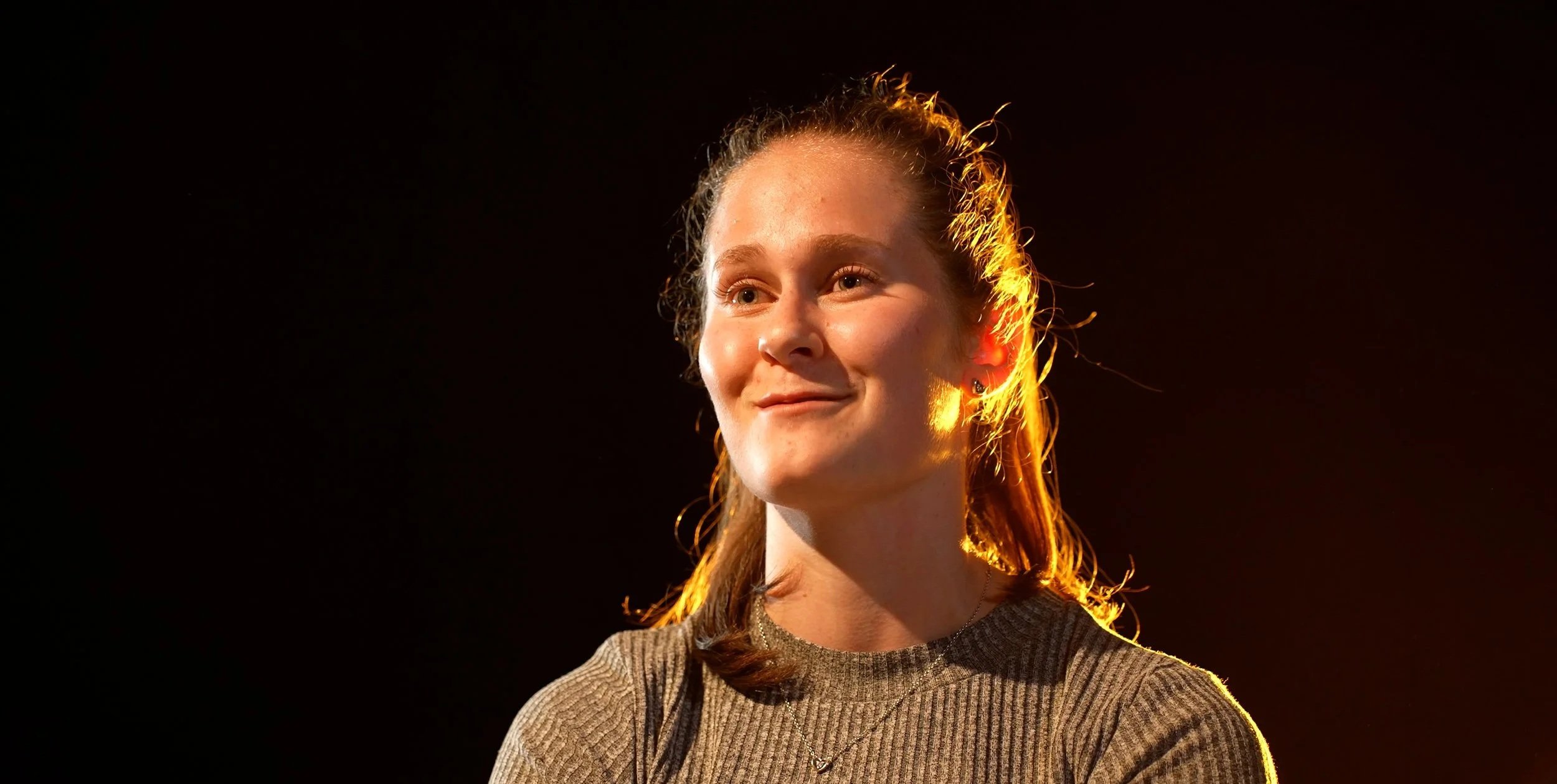 A young woman with red hair and earrings, smiling against a black background, illuminated by warm lighting.