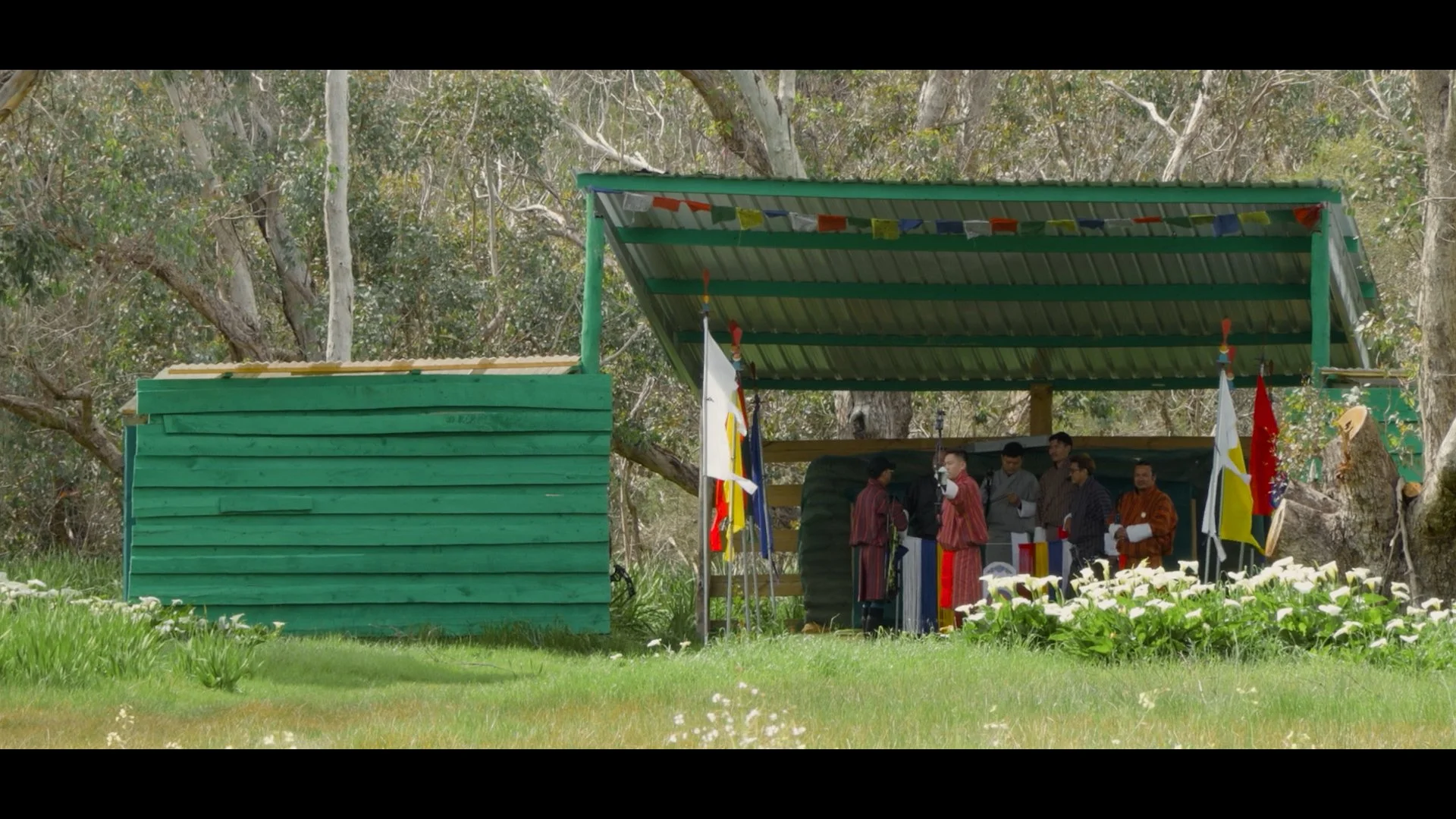 A group of people dressed in traditional Bhutanese clothing gathered under a green metal-roofed shelter decorated with colorful prayer flags, set in a wooded outdoor area.