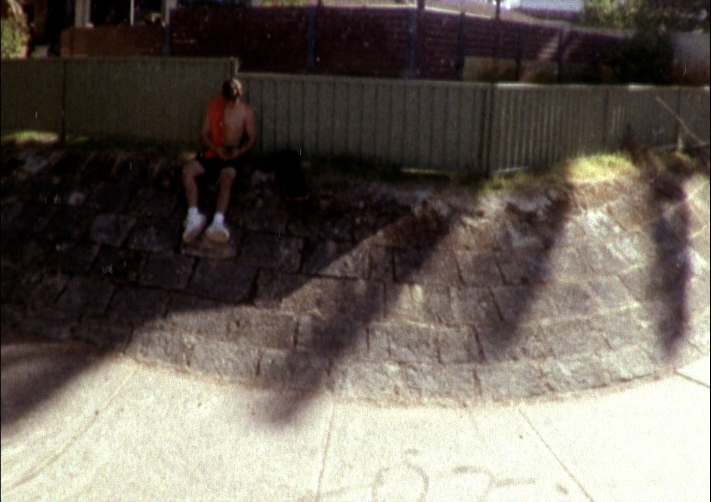 A person sitting on a stone ledge outdoors, shirtless, wearing shorts and white shoes, near a wooden fence at night.
