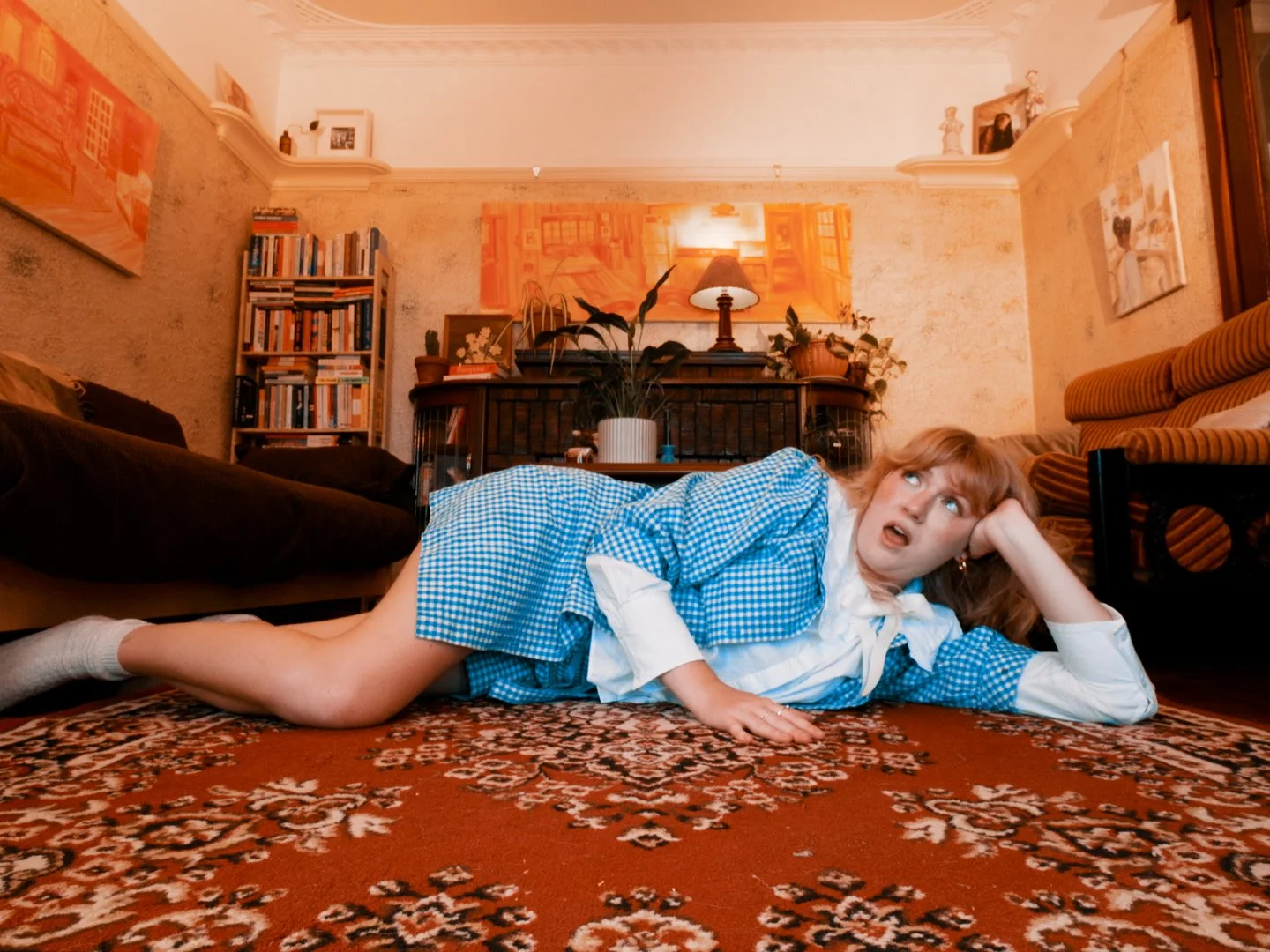 A woman lying on her side on a patterned carpet in a living room, wearing a blue checkered dress and white shirt, with a surprised or confused expression on her face.