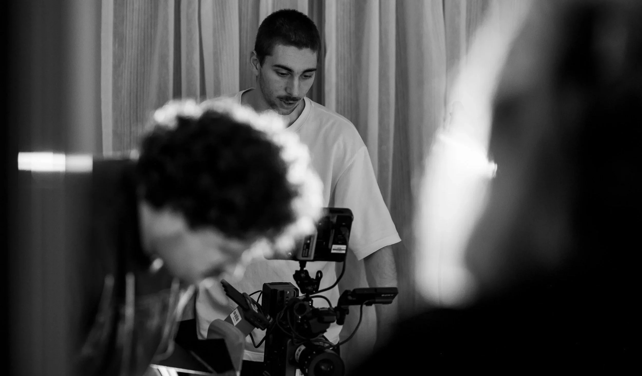 A young man with short hair working with camera and filming equipment in a room with wood paneling, with blurred people in the foreground.