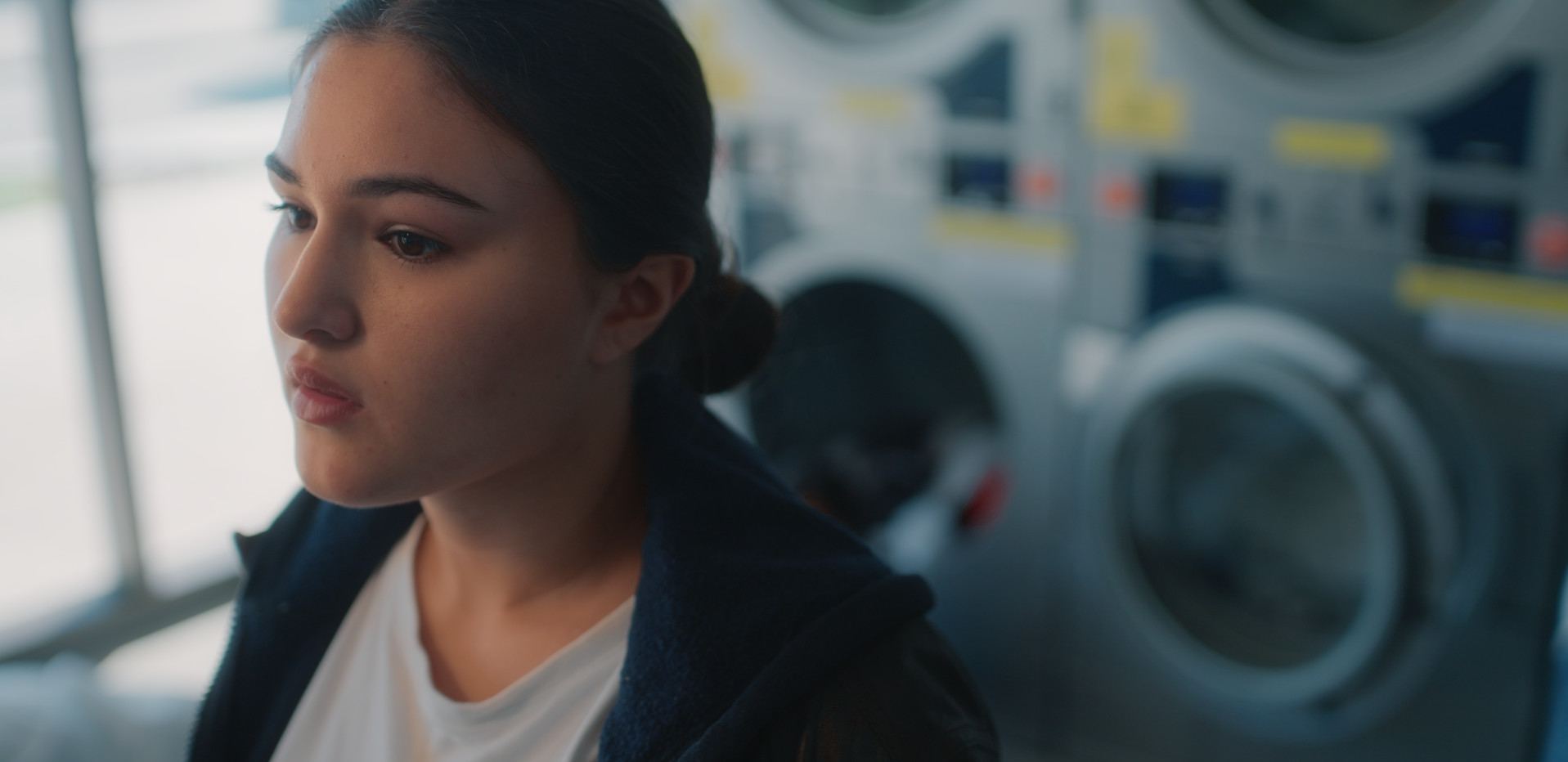 A young woman with dark hair tied back, wearing a dark jacket and white shirt, looking downward. She is in a laundromat with washing machines in the background.