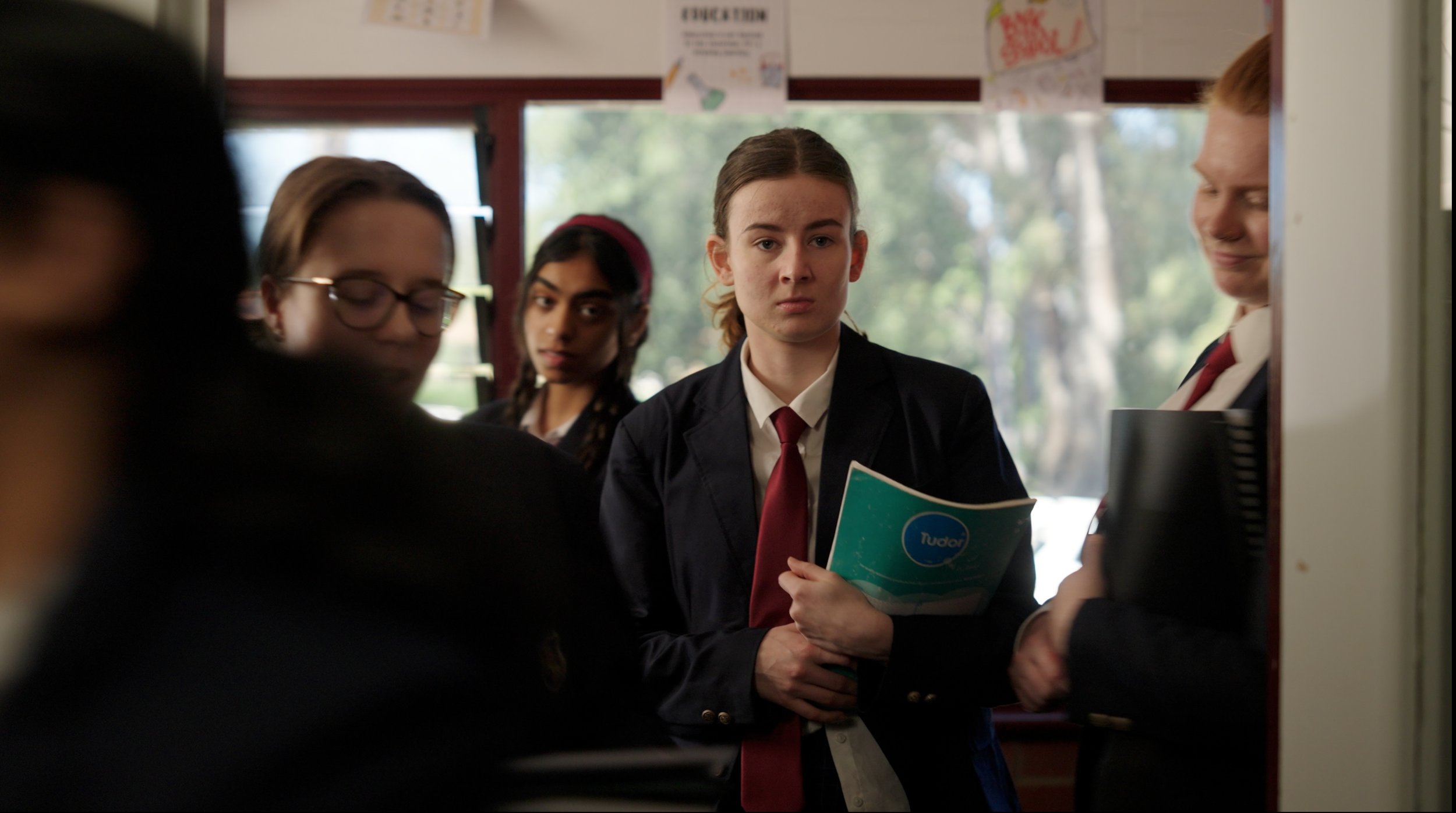 Group of students in school uniforms inside a school lobby or hallway, standing near a counter or reception desk, with large windows in the background.