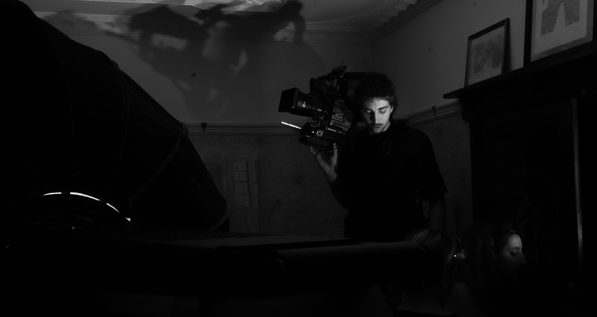 A person with dark hair operating a professional video camera indoors, with other people and framed pictures visible in the background.