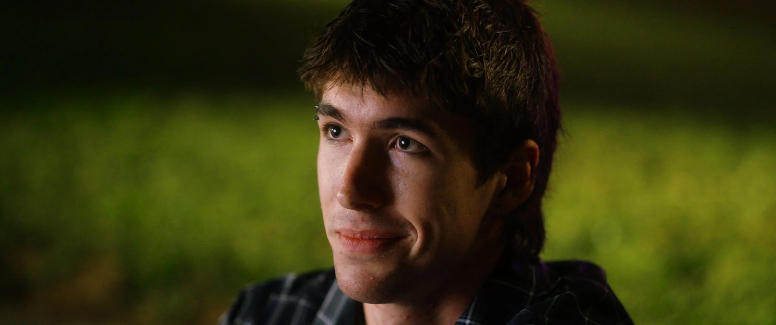 Close-up of a young man with brown hair, light skin, and blue eyes, looking slightly to the left, outdoors at night with blurred greenery in the background.