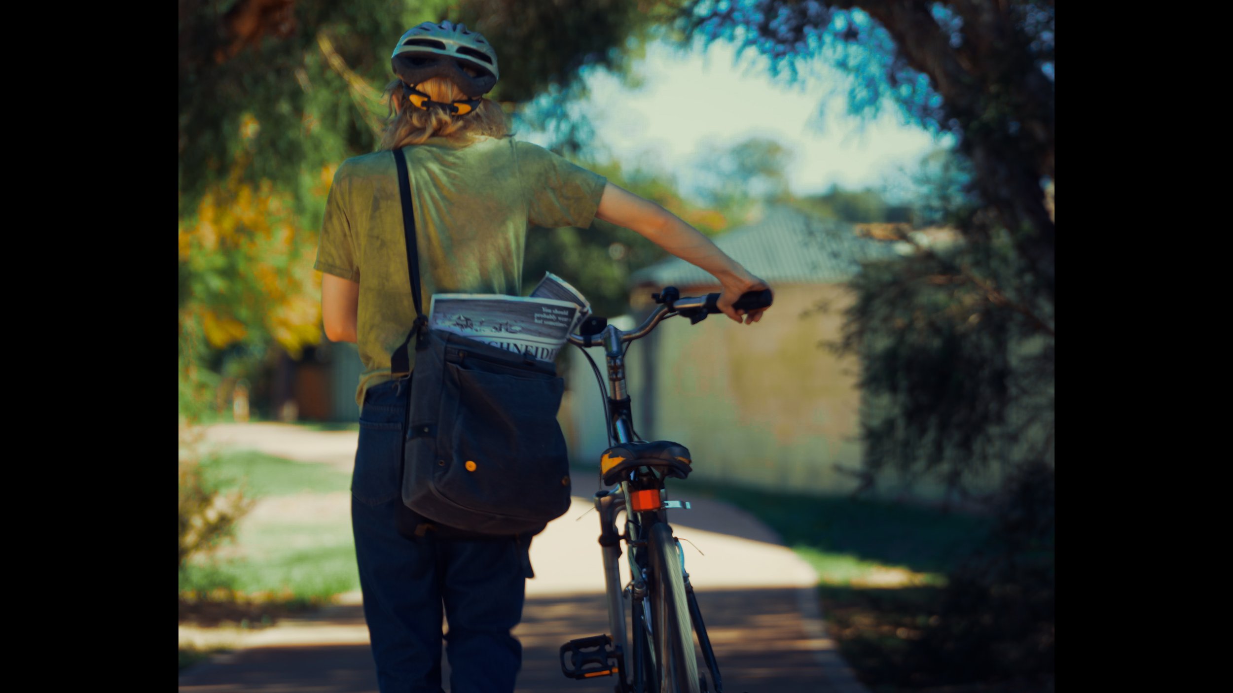 Person walking with a bicycle, wearing a helmet and carrying a bag with a magazine, on a sidewalk in a residential neighborhood with trees.