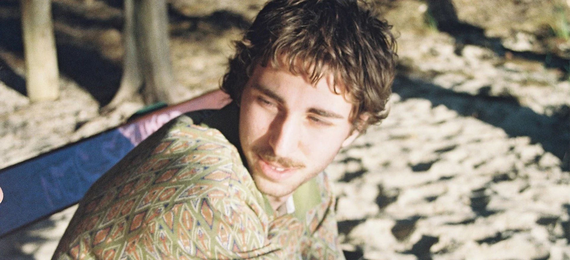 A young man with curly brown hair and a beard, wearing a colorful patterned jacket, looks to the side outdoors on a sandy beach with wooden structures in the background.