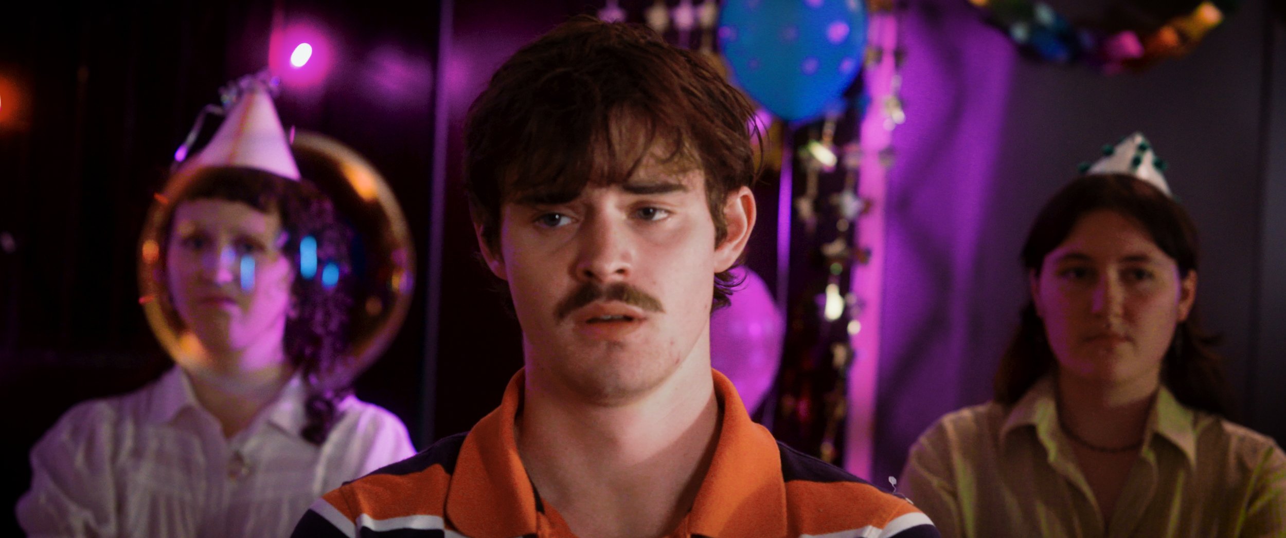 A young man with a mustache and wavy hair looking confused or surprised at a birthday party with party hats, surrounded by colorful decorations and purple lighting.
