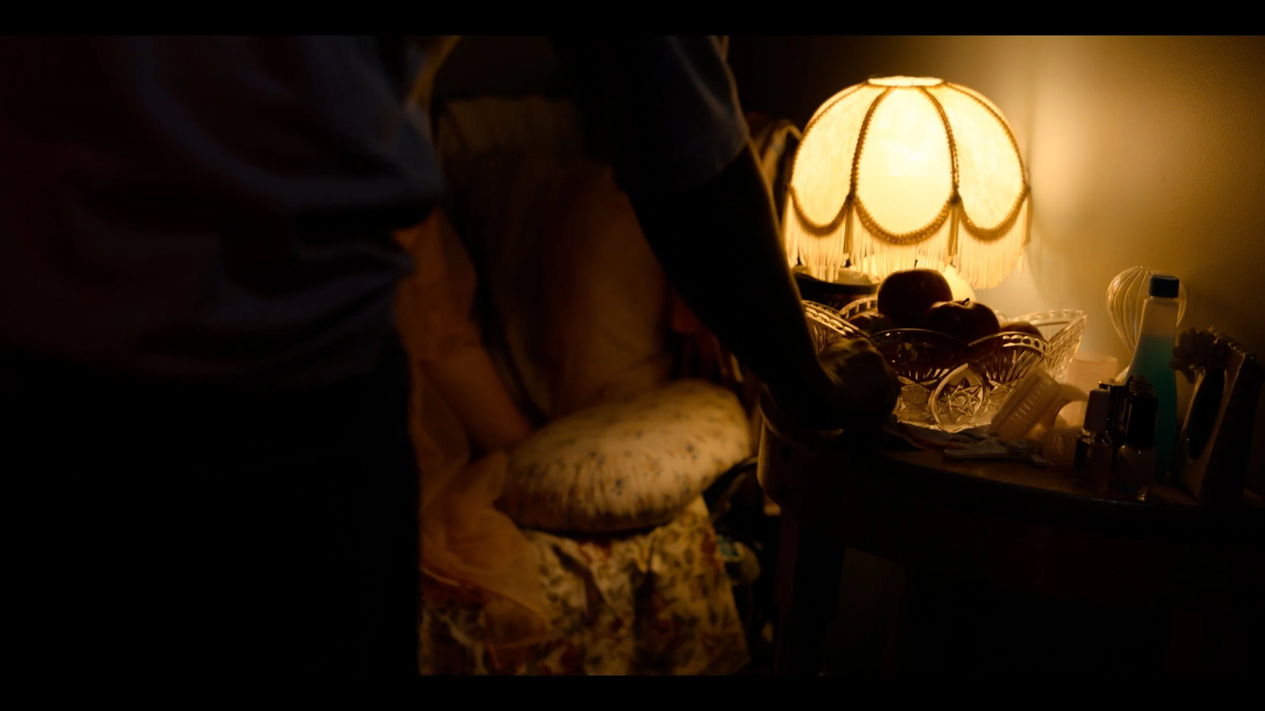 Dimly lit bedroom with a vintage lamp on a nightstand, cluttered with various items including bottles, a small decorative basket, and a pillow on a floral bedspread.