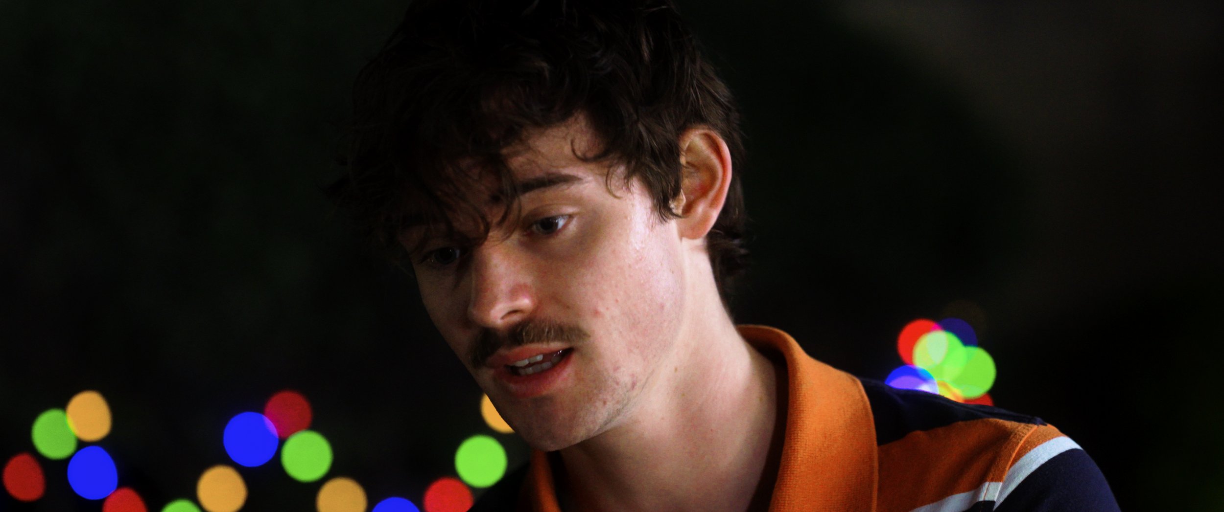 A young man with wavy brown hair and a light mustache is looking down and slightly to his left. He has pale skin with some blemishes and is wearing an orange and black shirt with stripes. The background features colorful out-of-focus lights, primarily red, green, yellow, and blue.