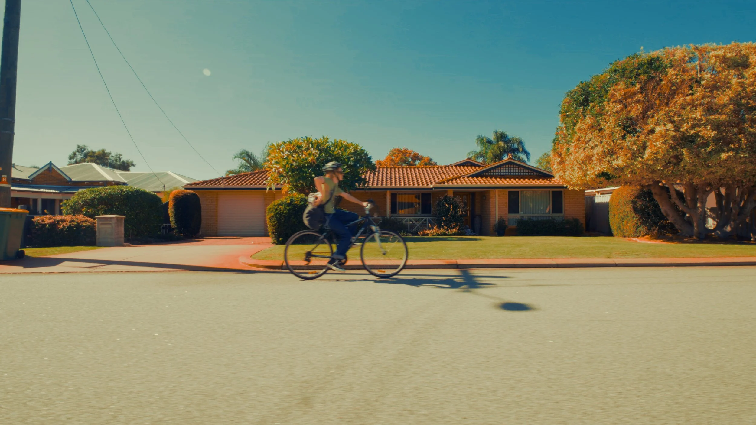 A person riding a bicycle on the street in front of a suburban house with a red tiled roof and well-maintained yard, trees with autumn-colored leaves, and a clear blue sky.
