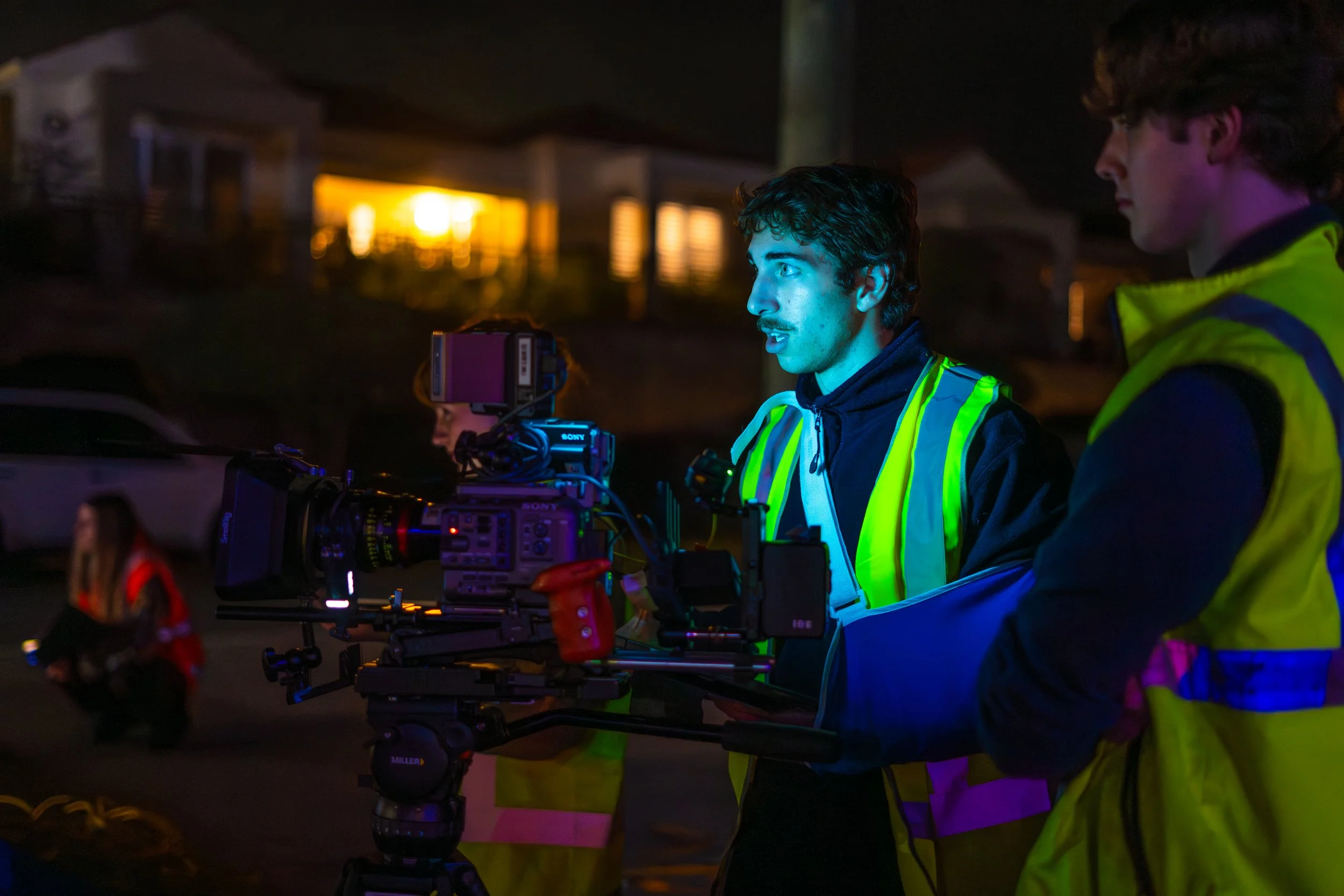 Two men wearing reflective safety vests operating a professional camera on a steady camera rig at night, with blurred background of a house and a woman in a red jacket in the foreground.
