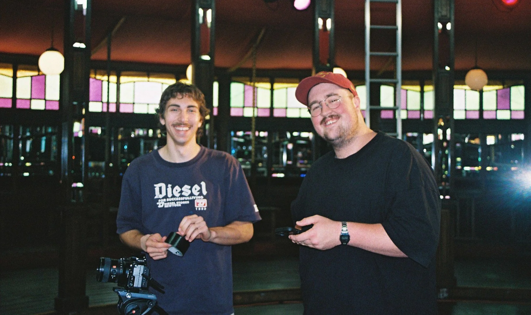 Two men smiling in a dimly lit indoor space with colorful stained glass windows in the background. One man wears a navy blue Diesel T-shirt and holds a small photo or card, the other wears a black T-shirt and red cap, holding a camera or phone.