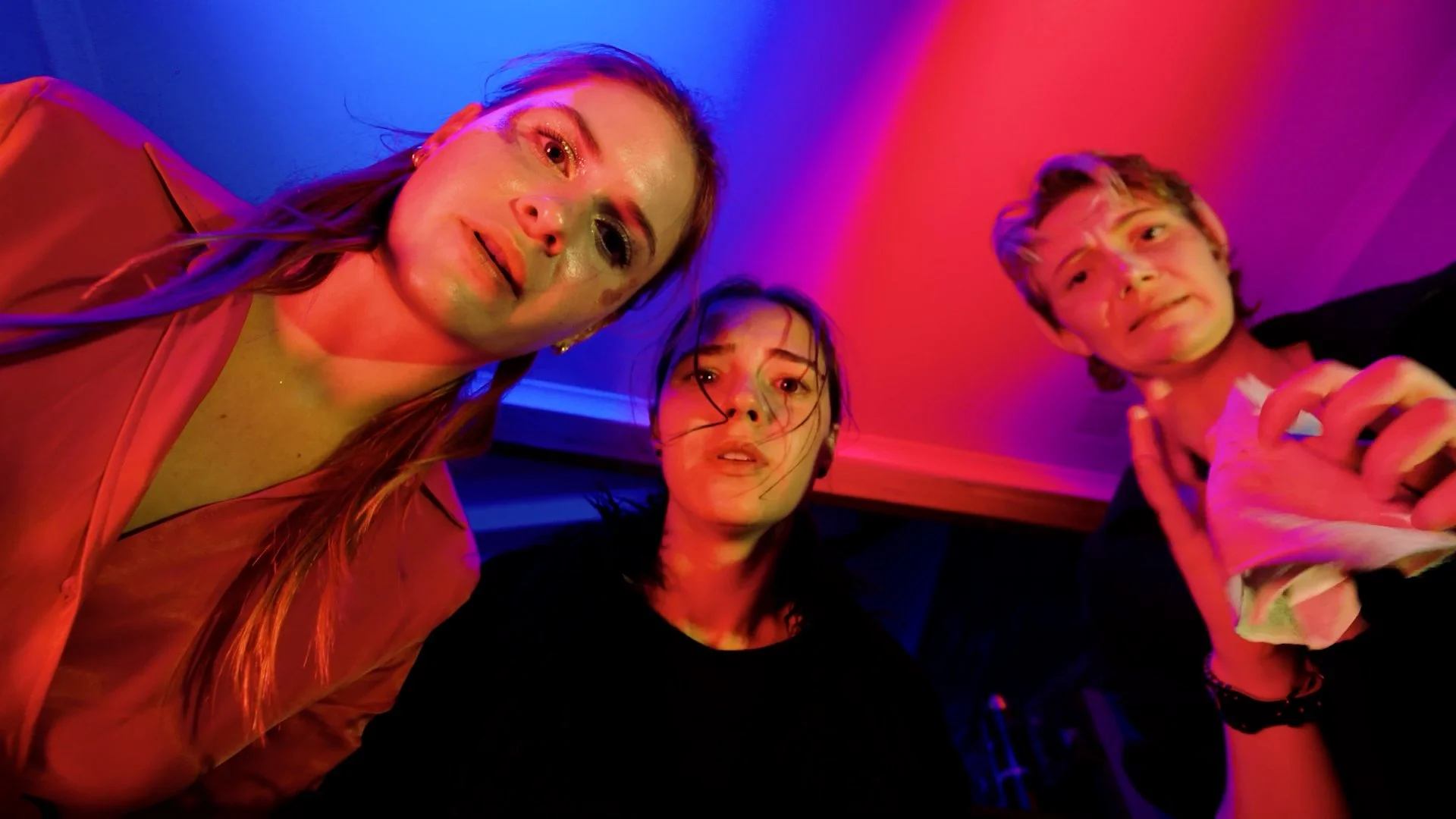 Three young women and one young man taking a selfie under colorful, neon lighting, looking up at the camera with relaxed expressions in a casual social setting.