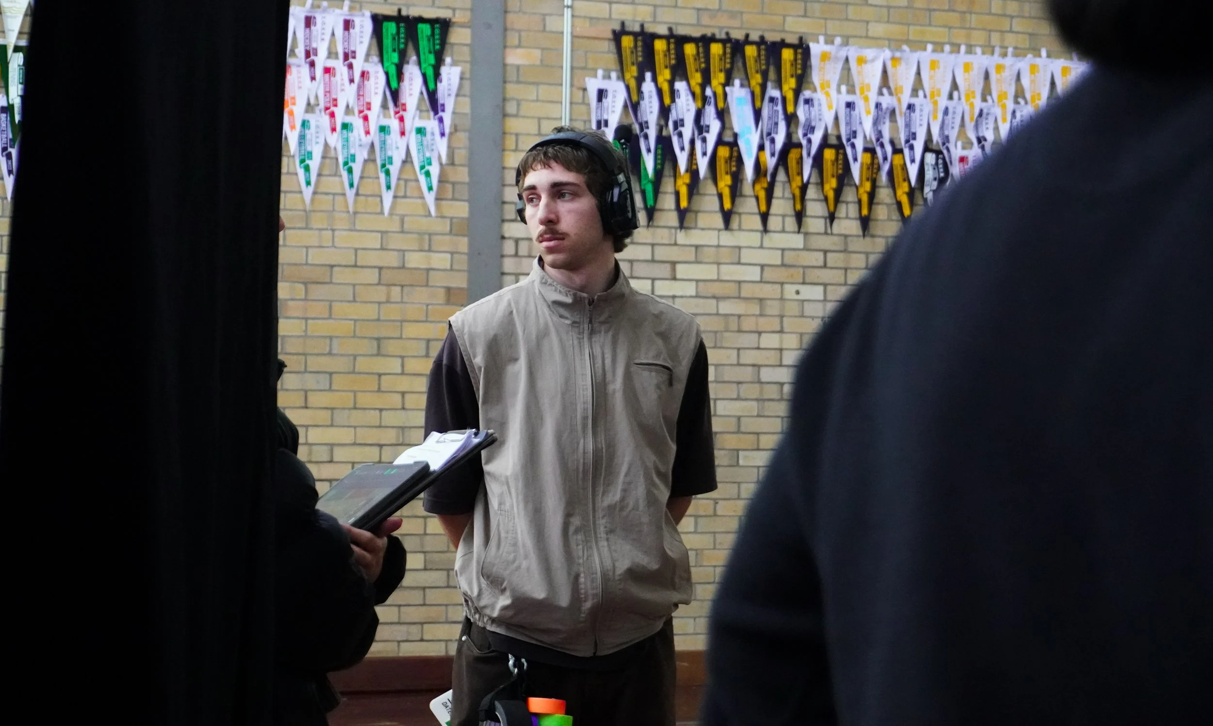 A young man with medium-length brown hair and a mustache, wearing a beige vest over a dark shirt, standing in front of a brick wall decorated with colorful banner flags. He has headphones on and is holding a clipboard, with people around him.