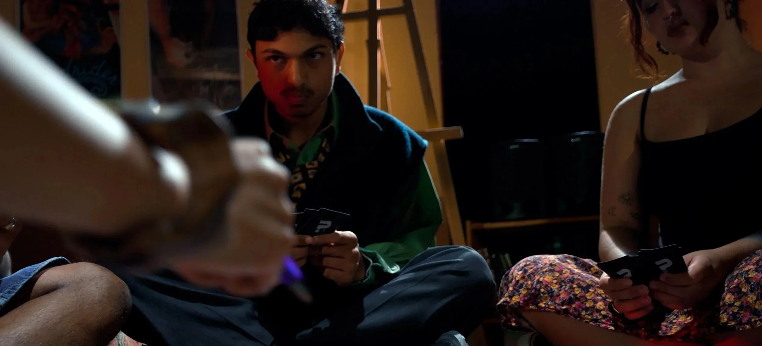 Two people sitting on the floor, the man looking forward with a neutral expression and the woman holding a card, both in a dimly lit room.