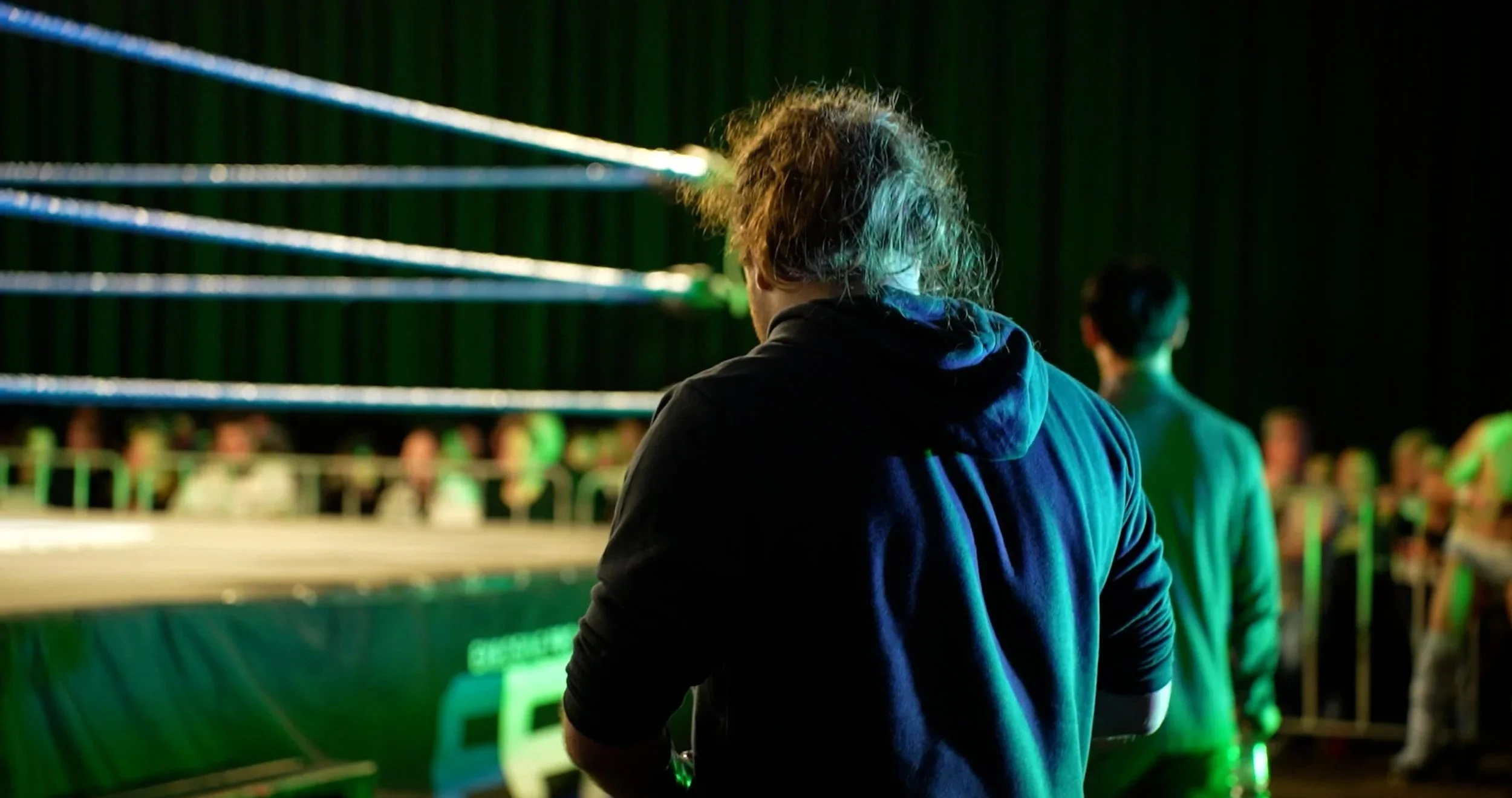 A person with messy curly hair wearing a hoodie, standing outside a boxing ring and watching a match at night with green ambient lighting.