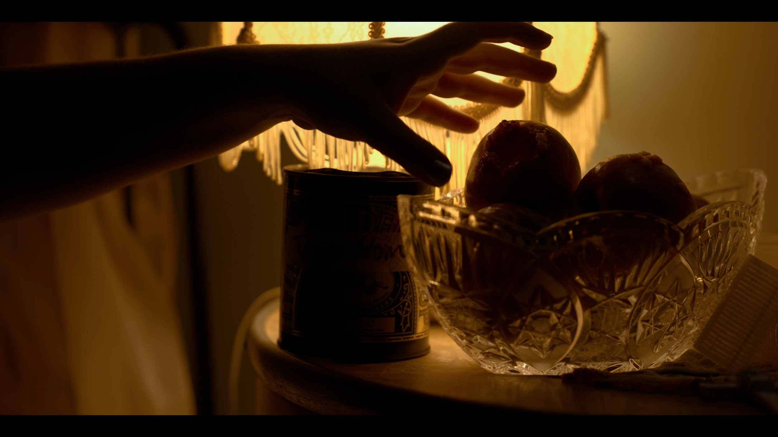 Silhouette of a hand reaching for fruit in a glass bowl on a kitchen counter, backlit by warm yellow lighting from a lamp with a lace shade.