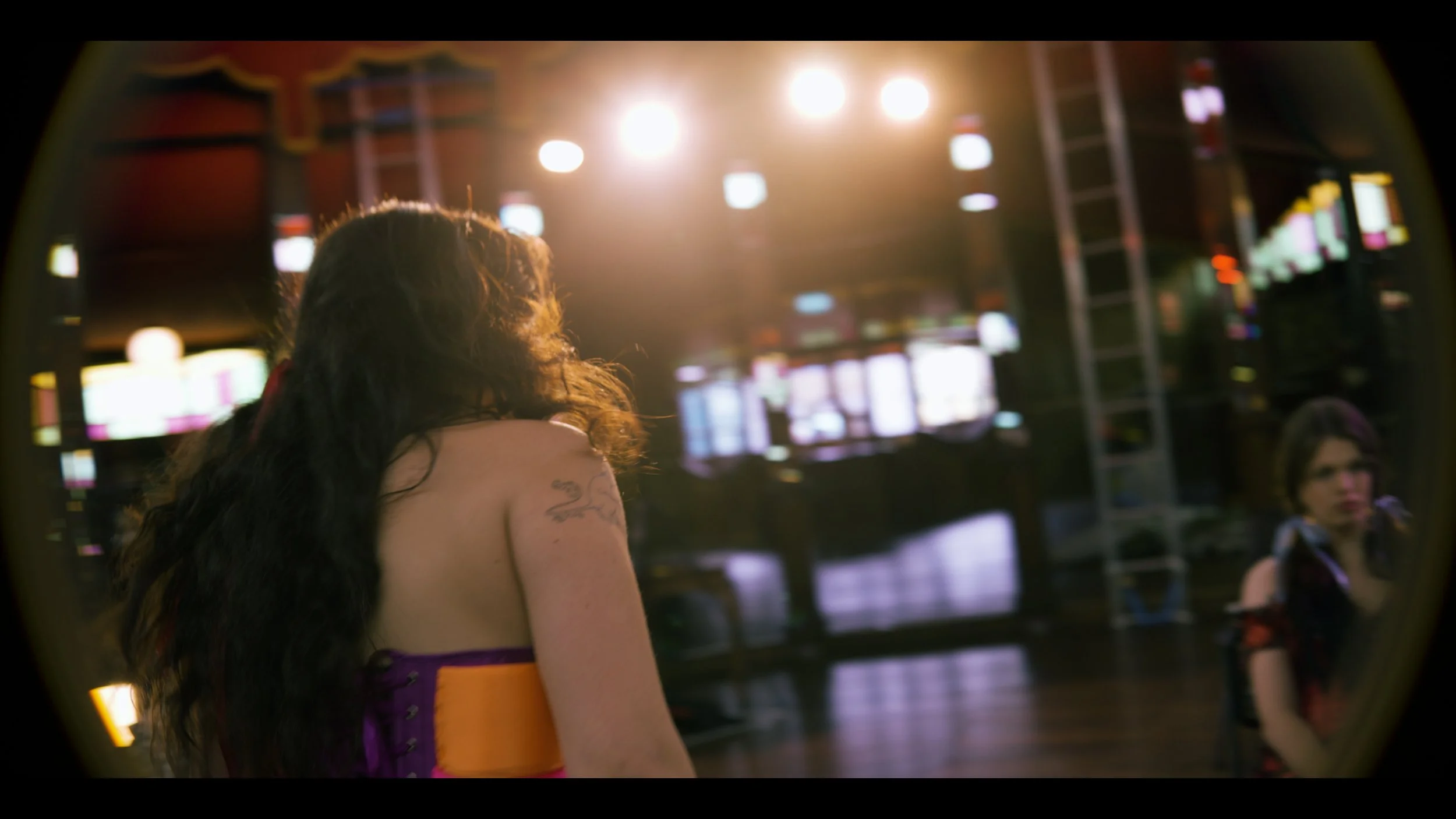 A woman with curly hair and a tattoo on her arm, wearing a colorful top, standing in a dimly lit area with bright lights and digital screens in the background.