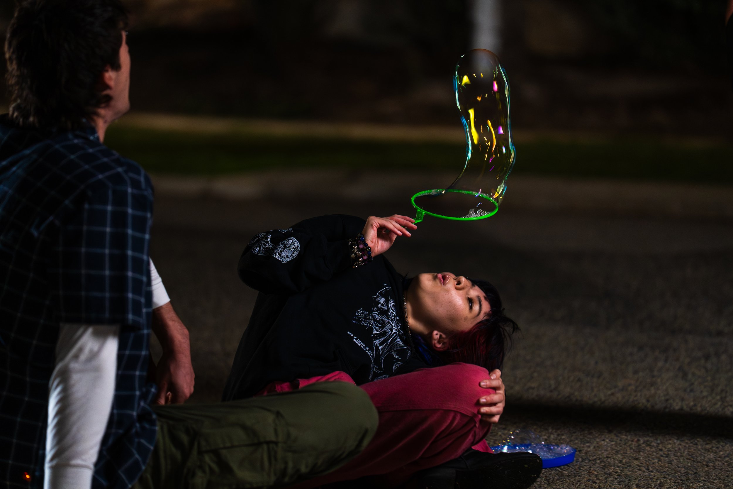 A group of four people sitting outdoors at night, one woman lying back and holding a lit soap bubble, while the others watch. One person is standing, another sitting on the ground, and one lying back with her arm around another woman.