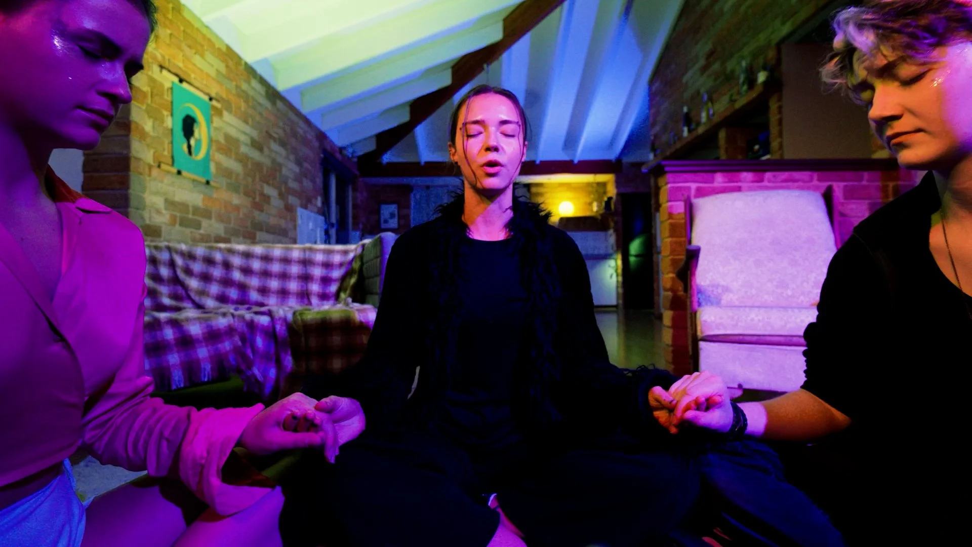 Three women sitting on the floor with eyes closed, holding hands in a prayer or meditation pose, inside a cozy room with brick walls and colorful lighting.