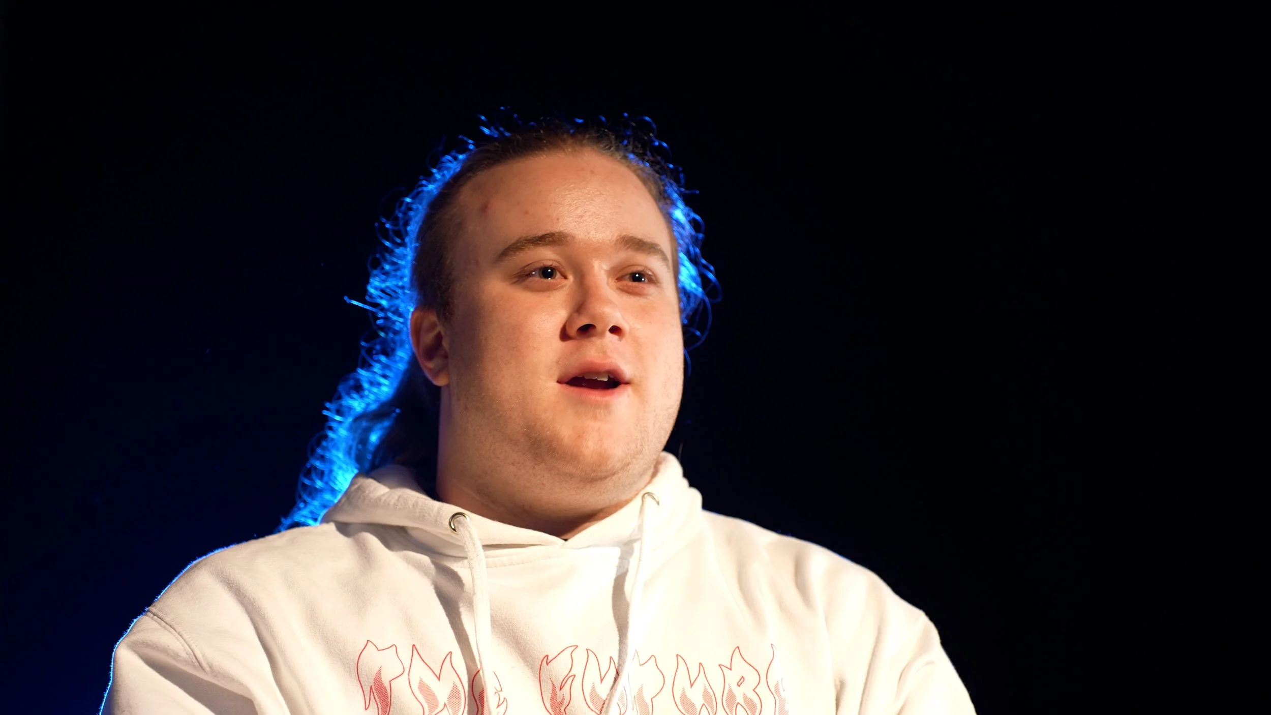 A young man with long hair and a surprised expression wearing a white hoodie against a black background, illuminated by blue lighting.