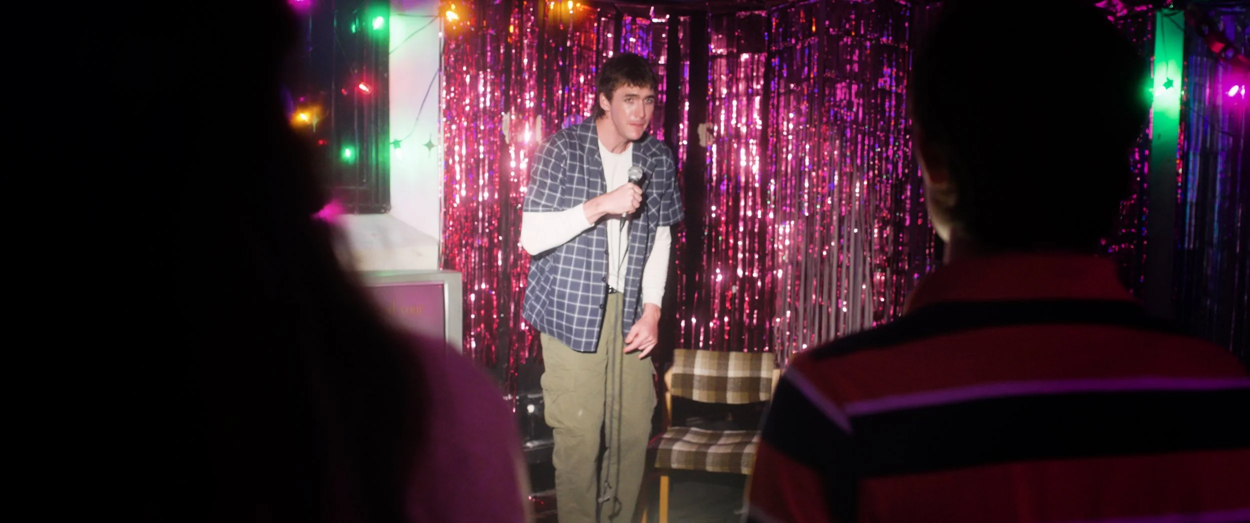 A young man performing stand-up comedy on stage with a microphone in hand, set against a backdrop of pink and purple tinsel and colorful lights, with an audience watching.