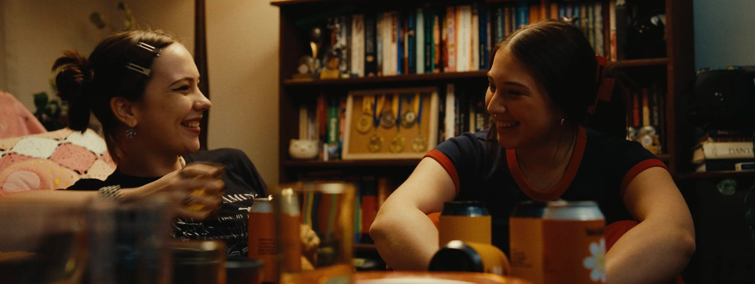 Two women sitting together at a table, smiling and laughing in a cozy room filled with books and collectibles, with cans and bottles on the table.