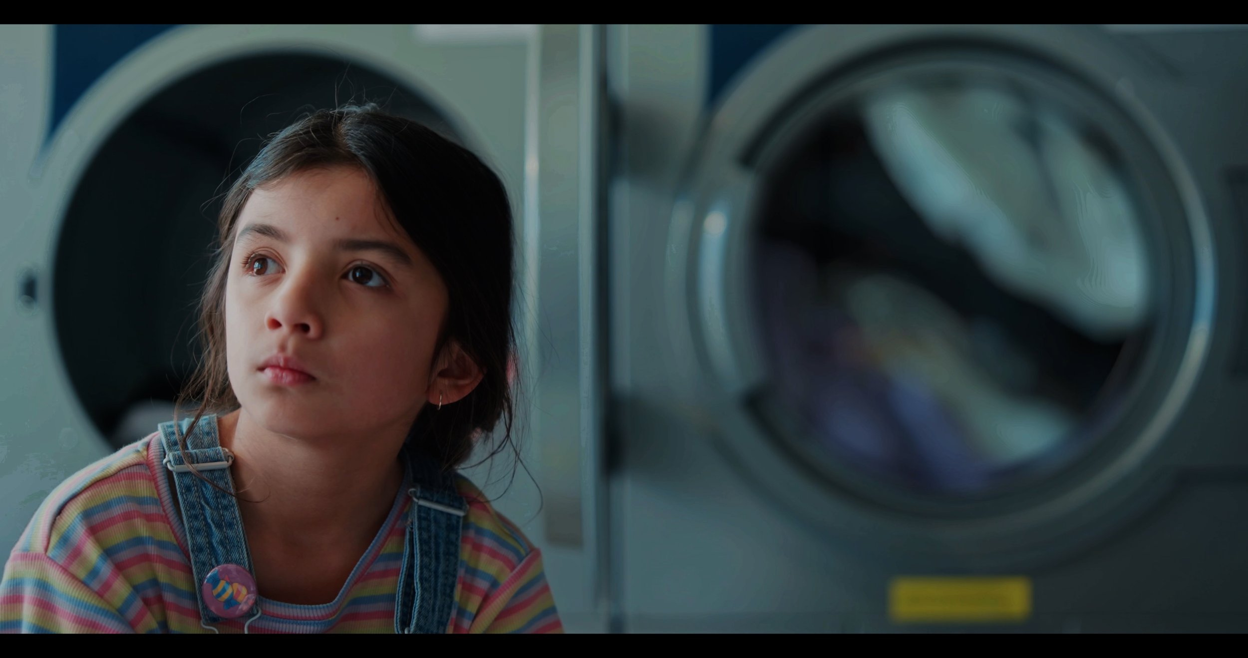 A young girl with dark hair and a striped shirt looking thoughtfully at washing machines in a laundromat.