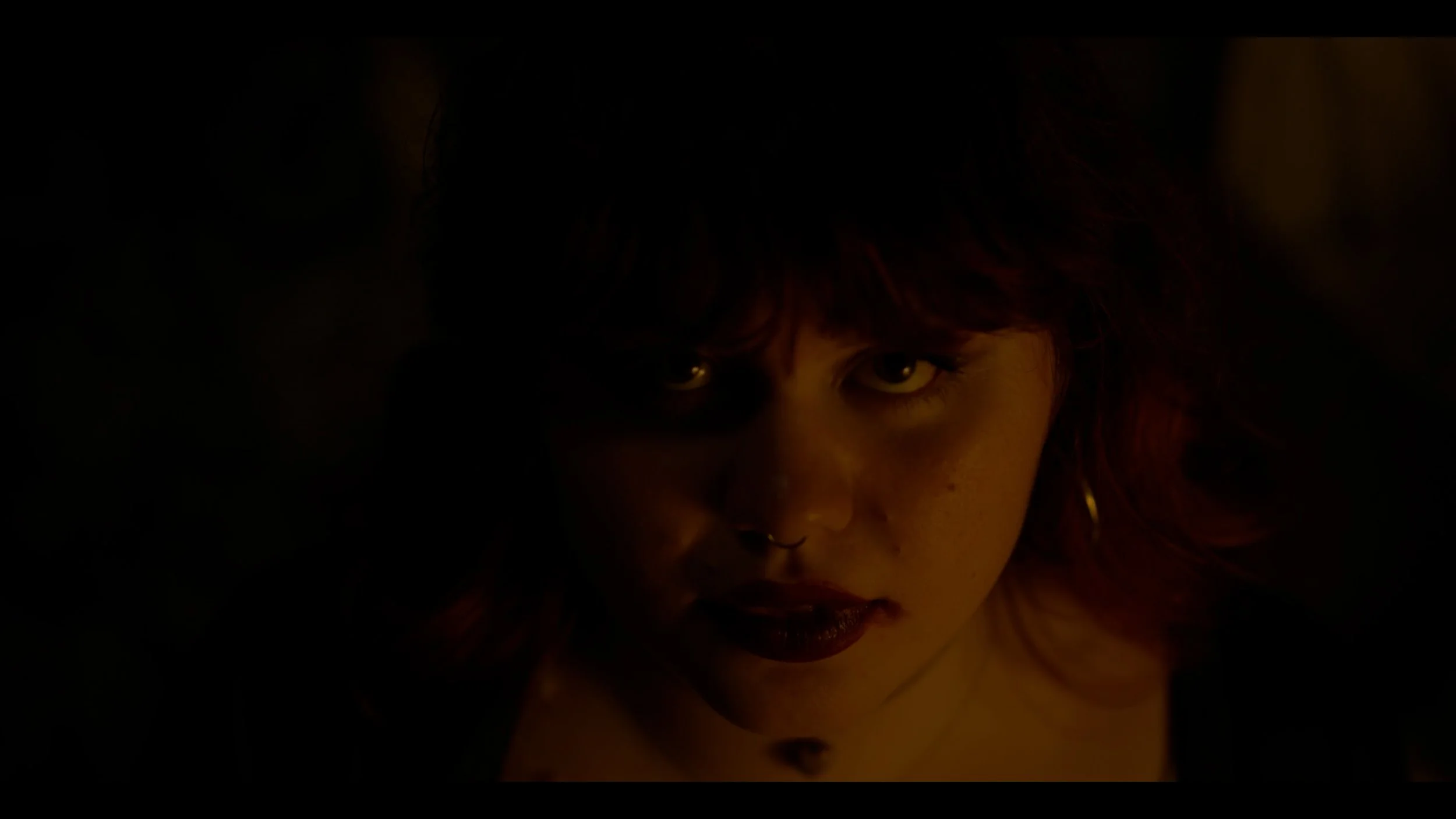 A person's face, partially obscured by shadows, with visible green eyes, dark hair with red highlights, and makeup including lipstick.