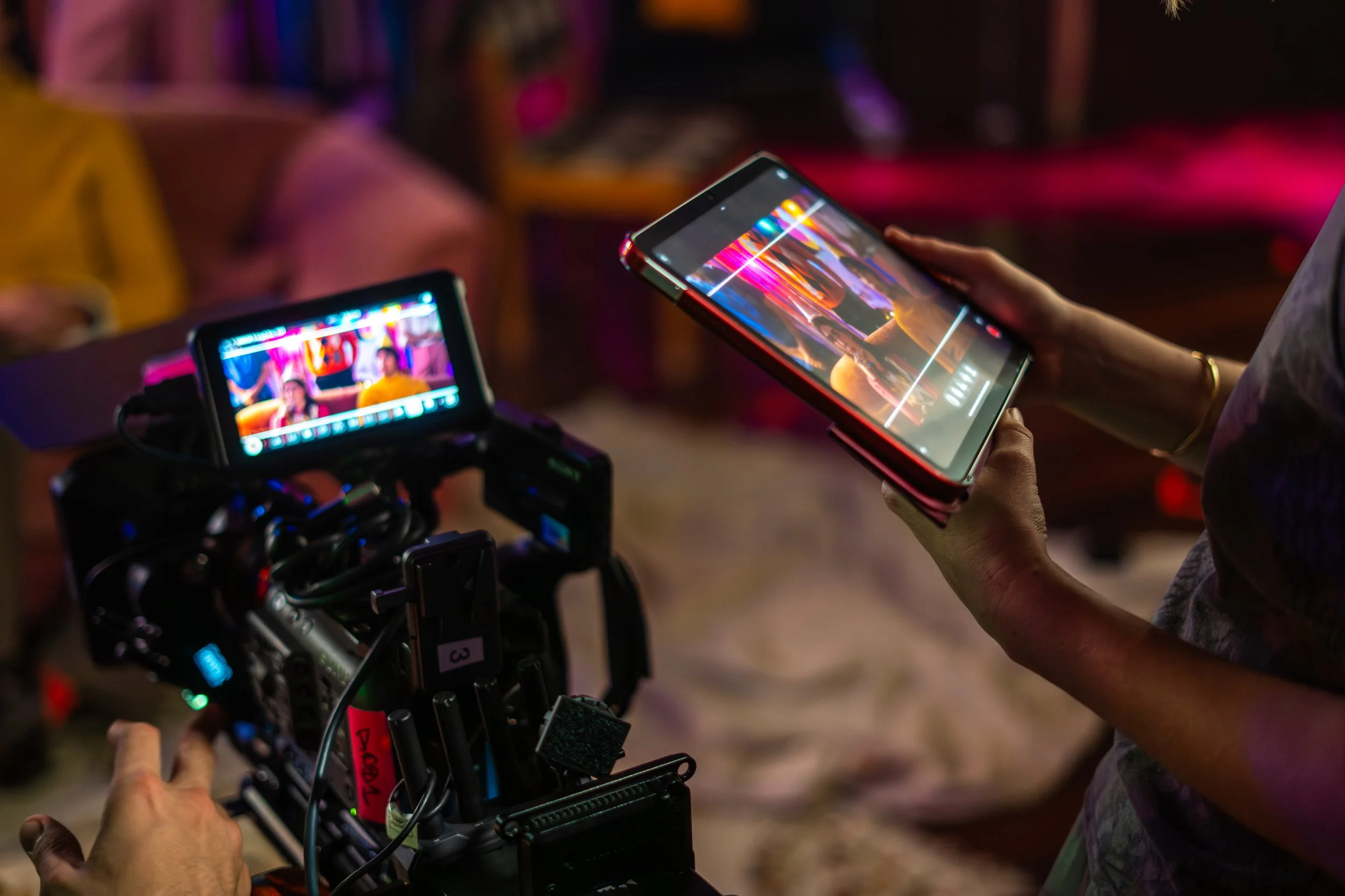 Person holding a smartphone with colorful display, a camera on a rig in the foreground, and a blurred background with vibrant lighting.
