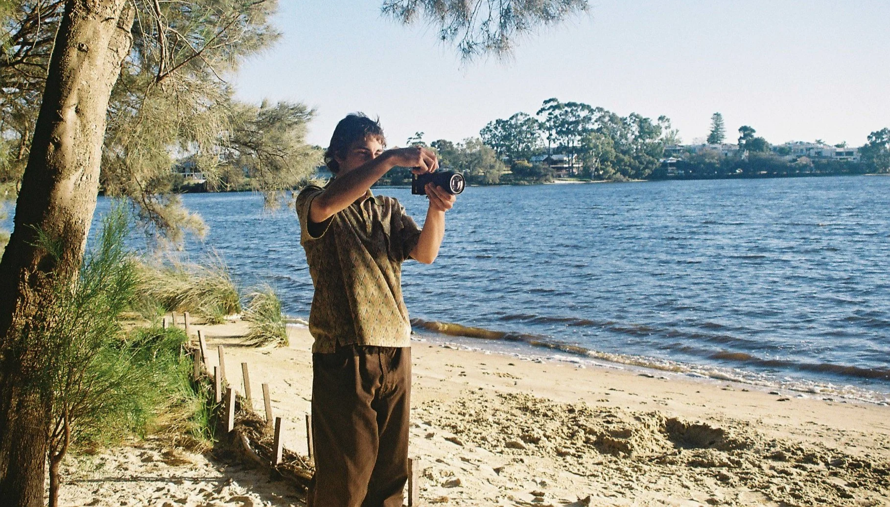 A person with short dark hair taking a photo or video with a camera on a sandy beach. They are wearing a patterned short-sleeved shirt and brown pants. Behind them are trees near a body of water under a clear sky.