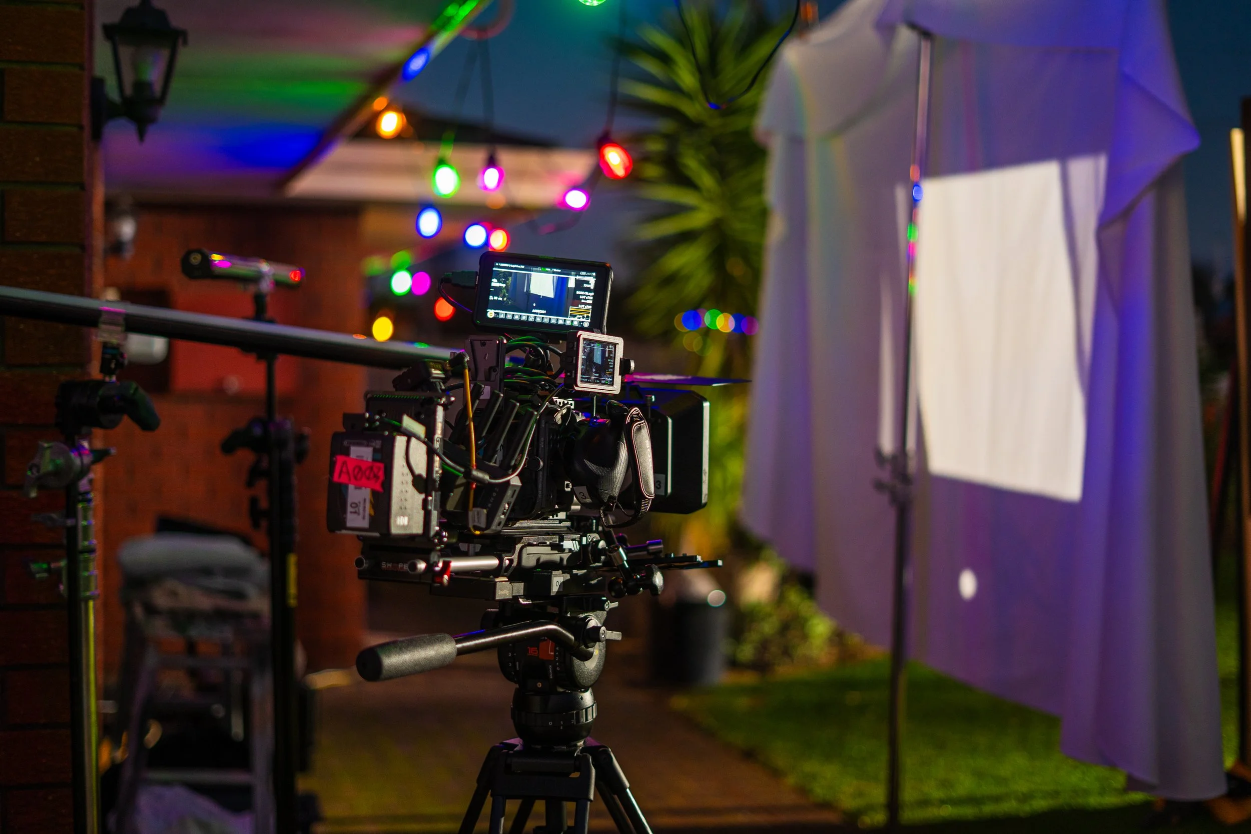A professional camera mounted on a tripod outdoors at night, with colorful string lights and a white screen or backdrop in the background.