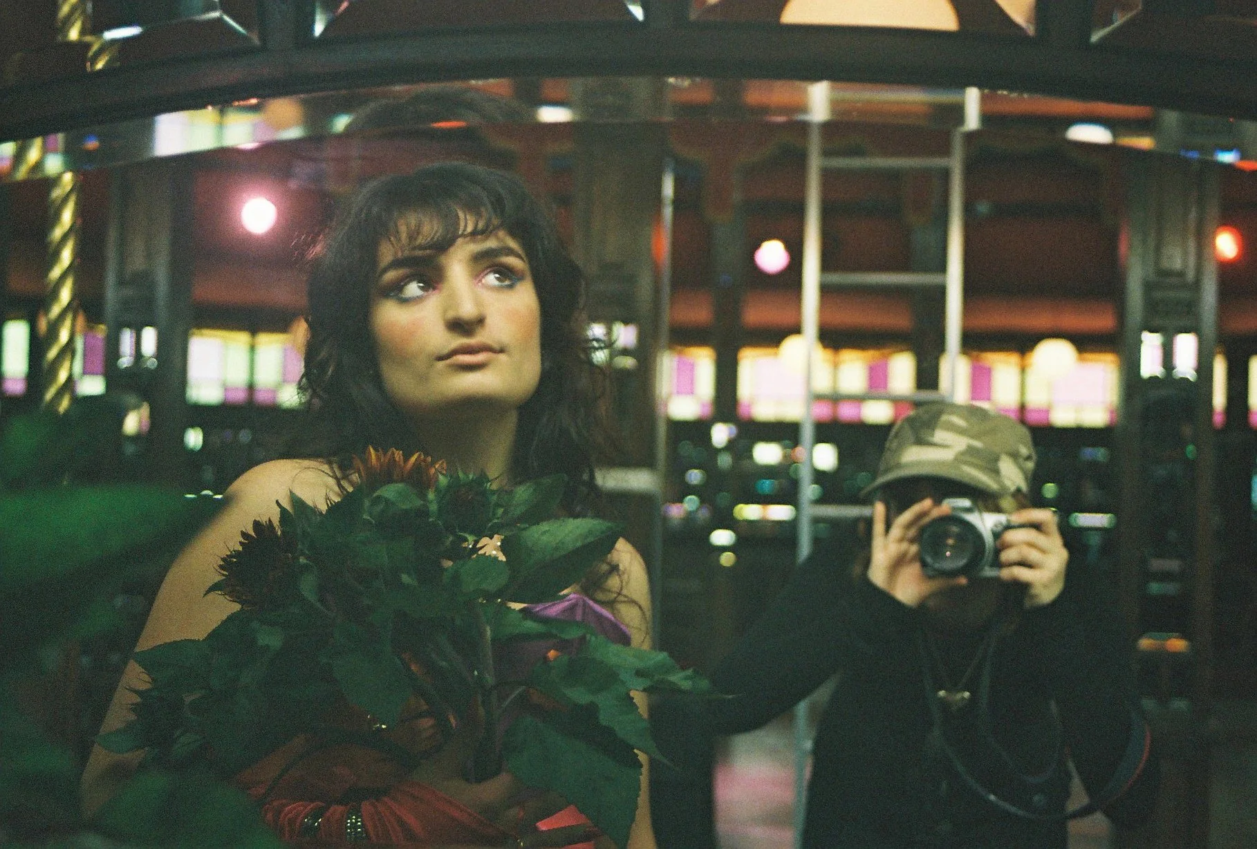 A person taking a mirror selfie in a carnival or fairground. The person has dark hair, a solemn expression, and is holding a bouquet of flowers. Another person, wearing a camouflage hat and dark clothing, is taking the photo with a camera. The background features colorful lights and carnival rides.