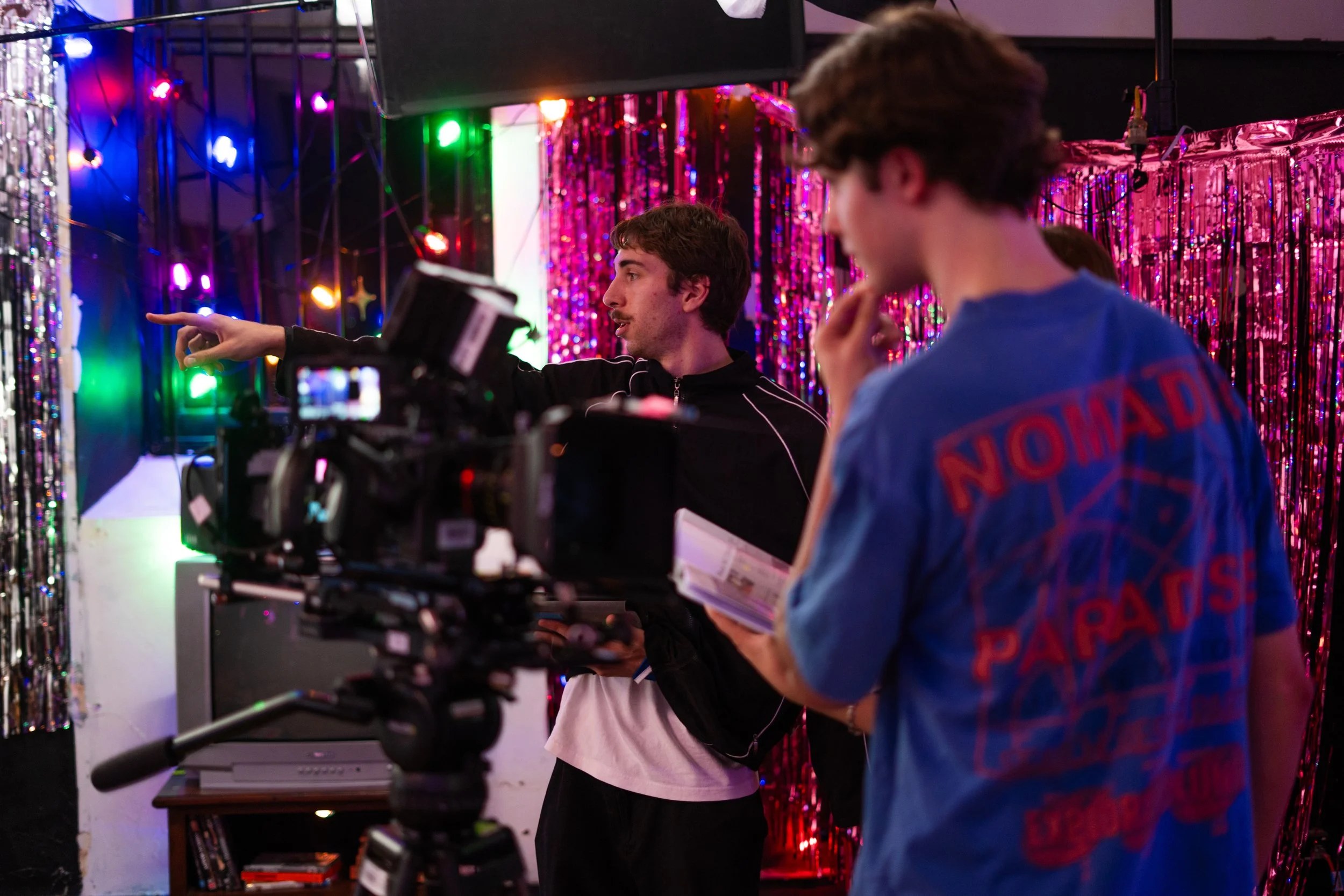 A man is filming with a professional camera while talking to a woman holding a script. They are in a colorful, festive setting with pink tinsel and multicolored lights.