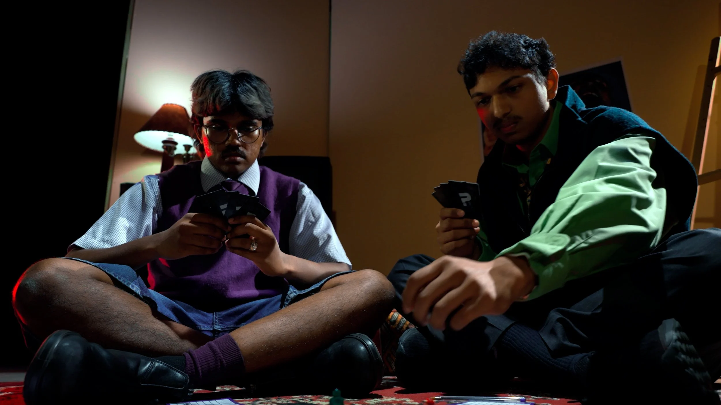 Two young men sitting cross-legged on the floor playing cards in a dimly lit room with a table lamp in the background.
