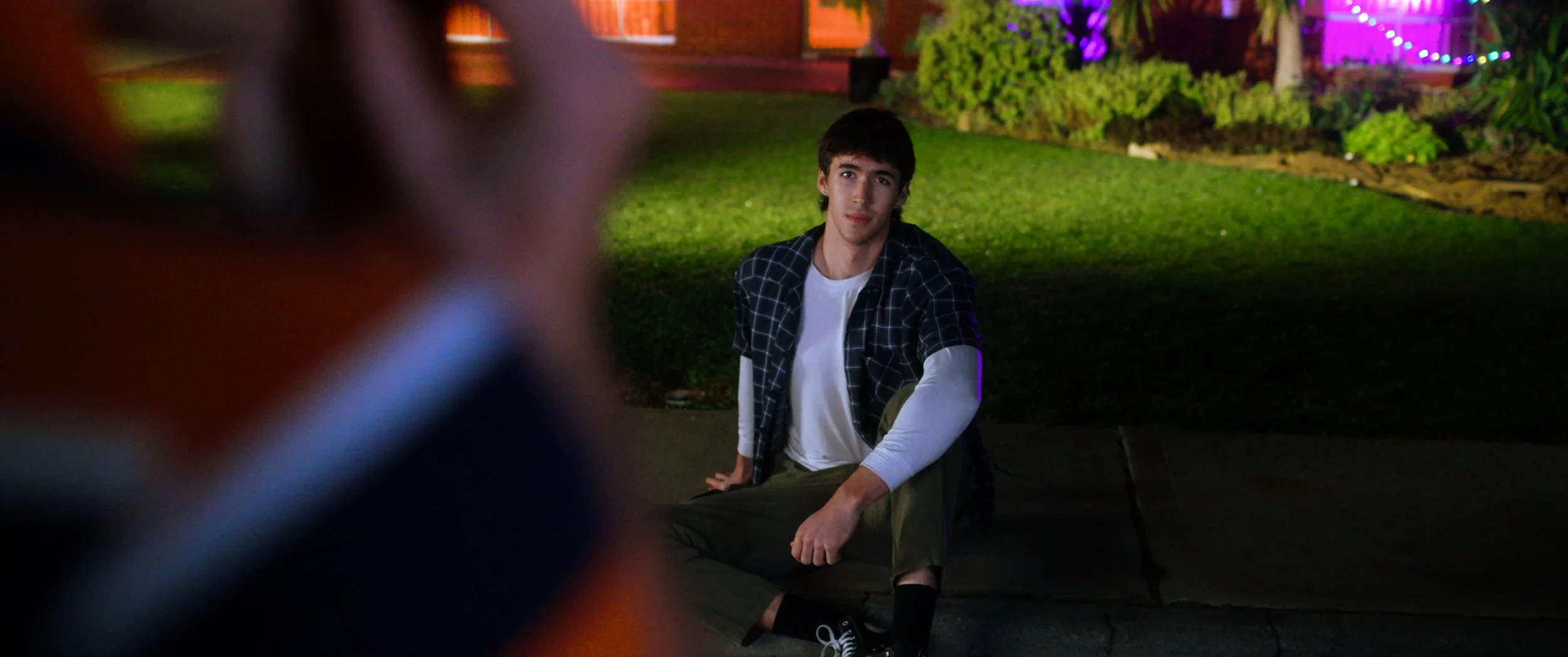 A young man with dark hair wearing a black and white checkered shirt over a white long-sleeve shirt sitting on a low wall in a park at night with colorful lights and bushes in the background.