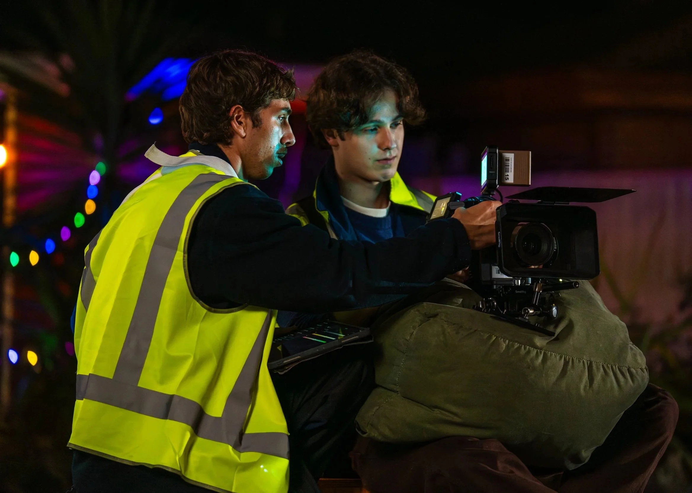 Two men, one wearing a bright yellow safety vest, are working together with a professional video camera at night, with colorful blurred lights in the background.