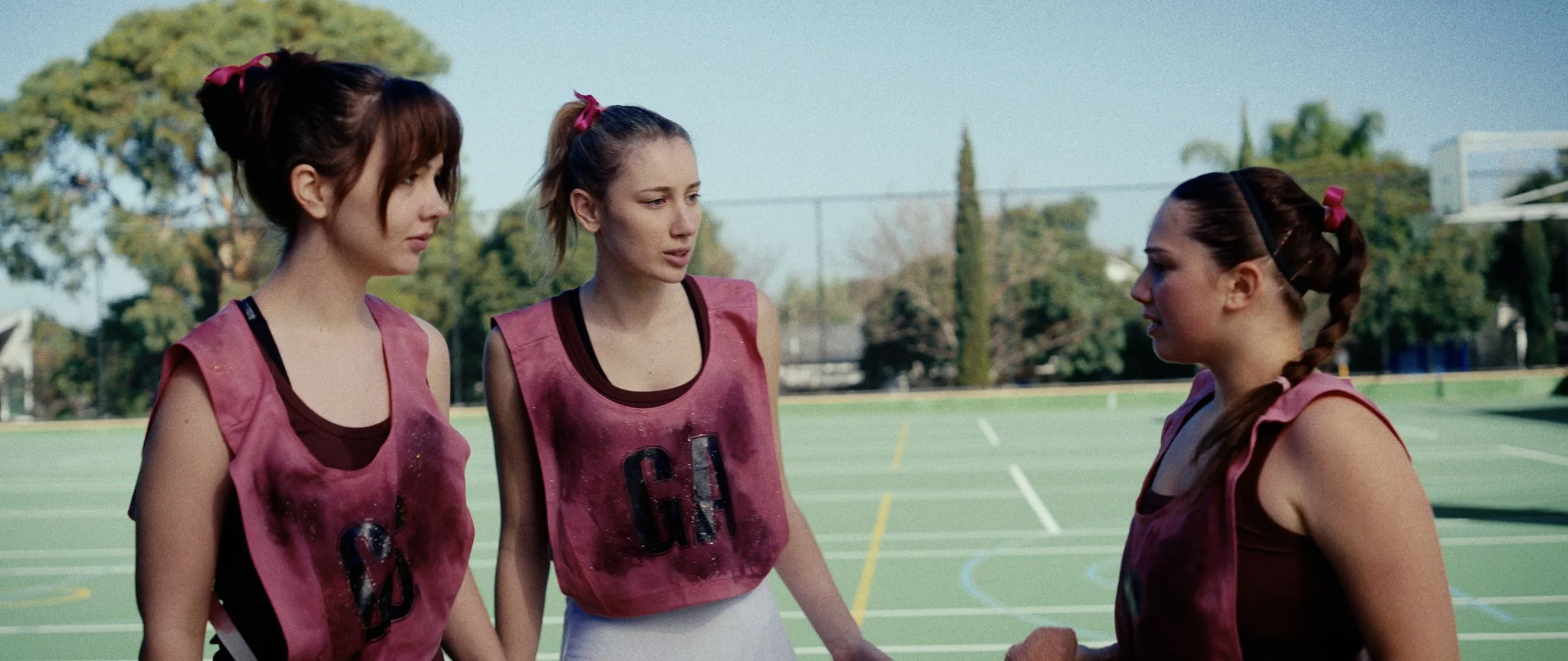 Three young women with athletic builds standing on a basketball court, wearing pink sports jerseys, having a conversation.