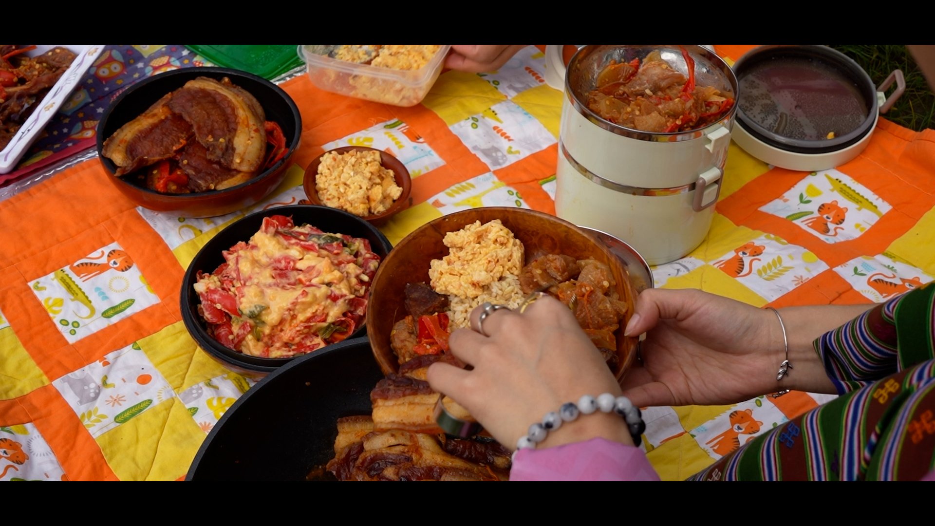 A colorful table with various homemade dishes including meats, rice, vegetables, and salad, with people serving themselves.