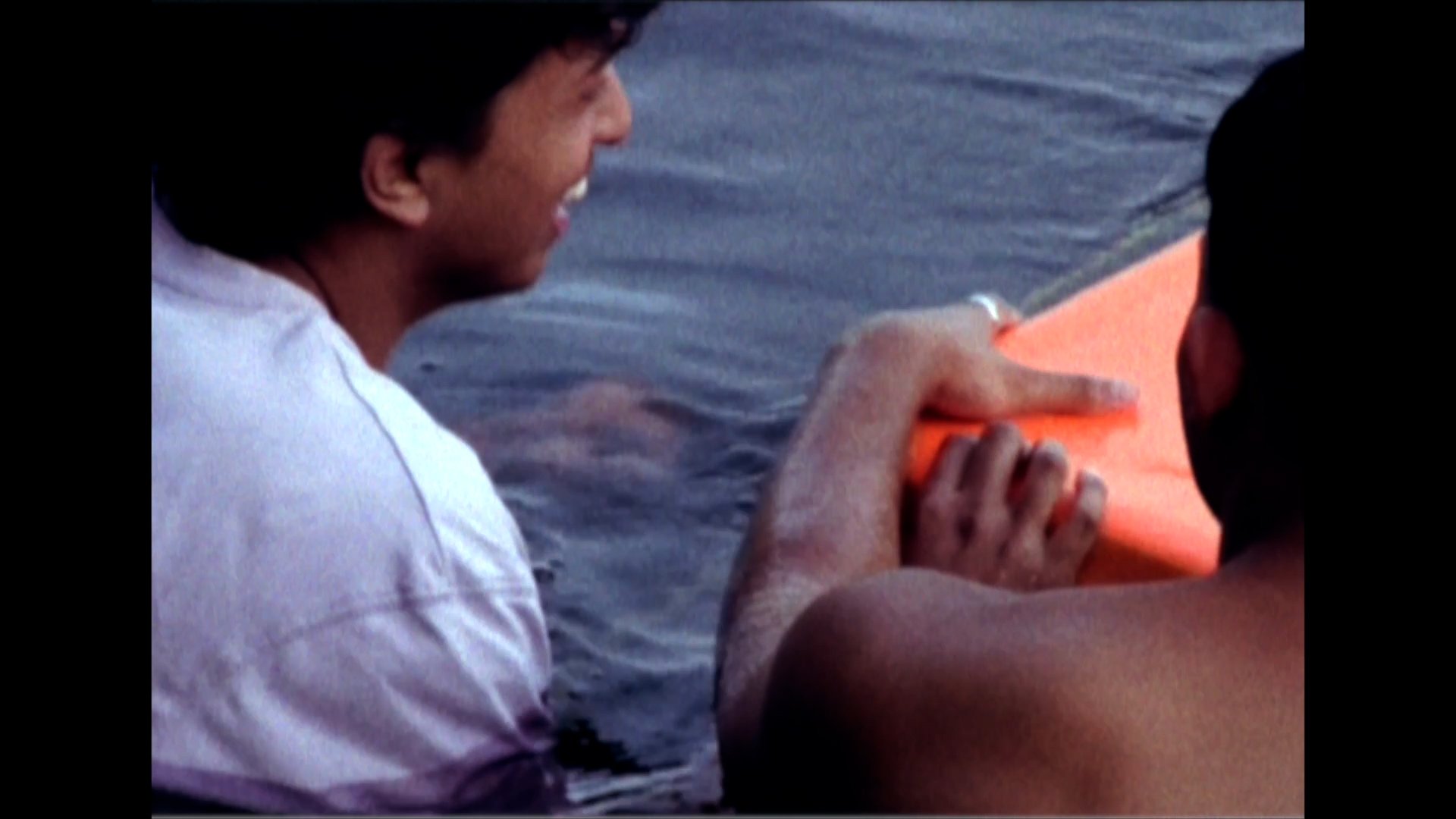 Two men sitting by the water, one holding an orange surfboard and the other resting.
