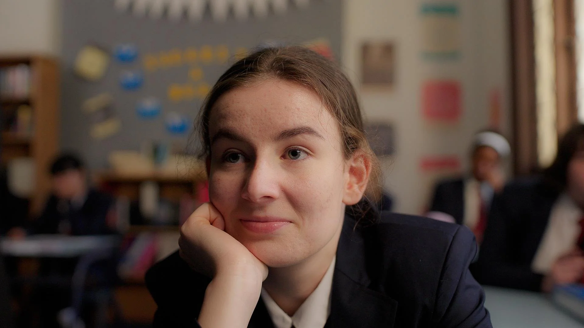 Close-up of a teenage girl with brown hair, blue eyes, and light skin resting her chin on her hand in a classroom setting. The background shows other students and educational decor.