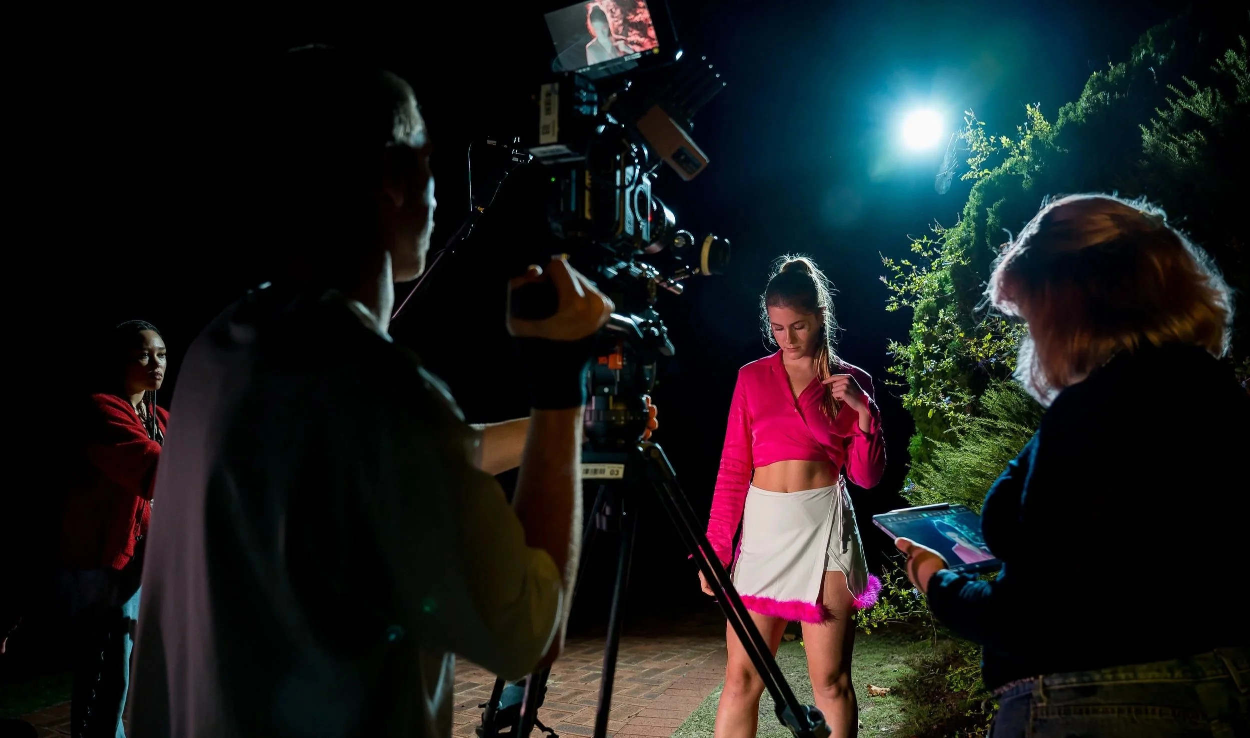 A nighttime behind-the-scenes photo of a music video shoot, featuring a female performer in pink and white clothing, being filmed by a camera crew with professional equipment, with trees and bright moon in the background.