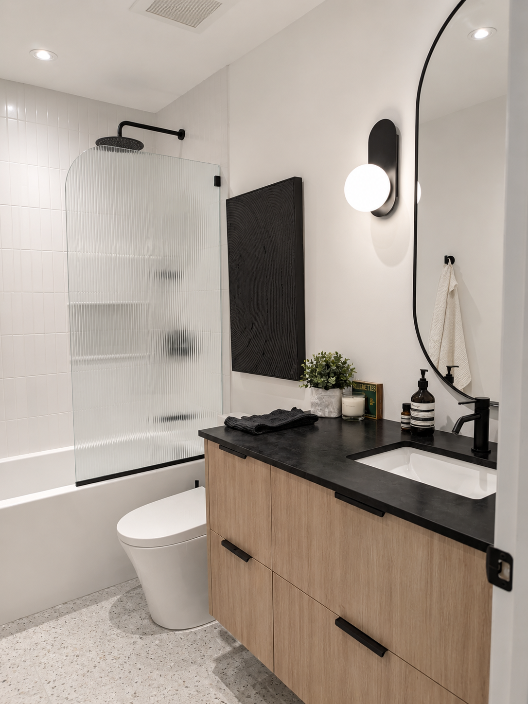 Modern bathroom with walk-in shower, black shower head, frosted glass divider, white toilet, wooden vanity with black countertop, oval mirror, black fixtures, wall-mounted lighting, towel, plants, candles, personal care products.